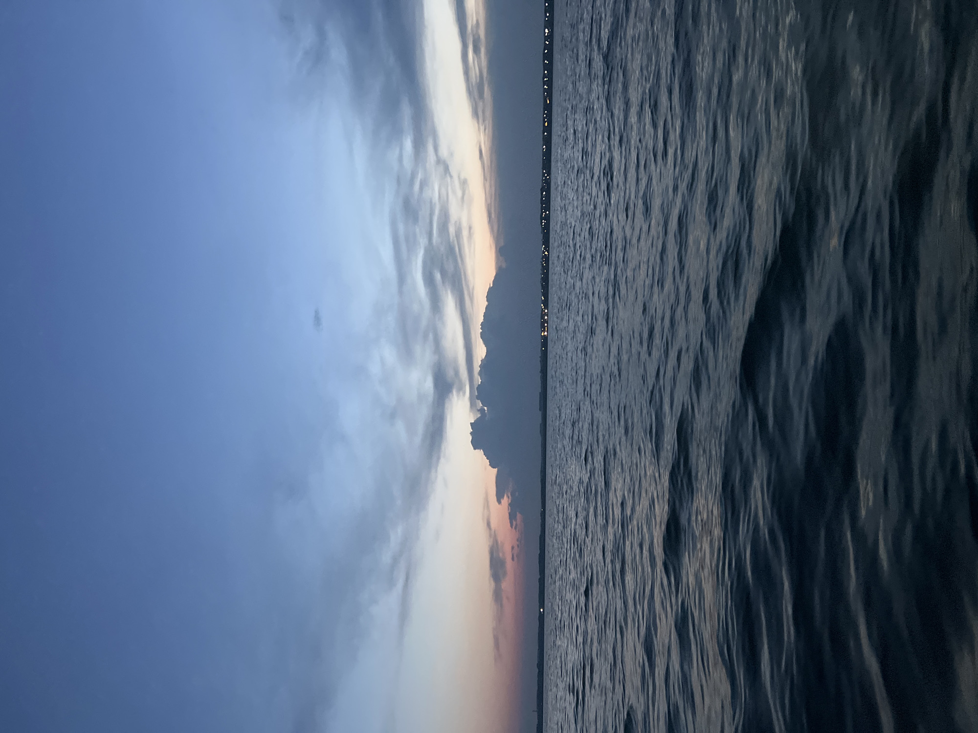 The image depicts a serene body of water, likely a lake or bay, with gentle ripples on its surface. The sky above is a canvas of twilight hues, transitioning from a deep blue at the top to softer shades of pink and orange near the horizon. Wispy clouds are scattered across the sky, adding texture and depth. In the distance, a faint line of lights suggests a cityscape or a coastal town. The overall mood is tranquil and atmospheric, evoking the quiet beauty of dusk. The location is the city of Novopichugovo, Russia.