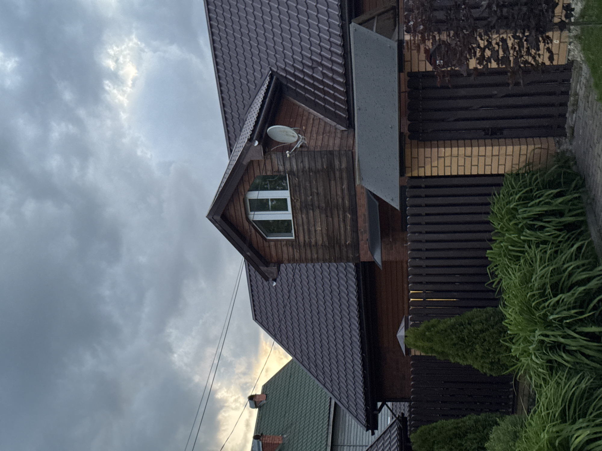 This media file captures a residential scene featuring a house and its immediate surroundings under an overcast sky. The image is framed to show the side of a two-story house, characterized by a dark brown tiled roof with a prominent dormer window on the upper level. The upper portion of the house is clad in dark brown wooden siding, where a small white satellite dish is mounted below the dormer. The window itself has white frames. Below the wooden siding, a section of light-colored brick is visible, partially obscured by a dark brown slatted fence.
In the foreground, lush green vegetation, including tall grassy plants and conical evergreen shrubs, grows along the fence line. To the left of the main house, another residential structure with a green tiled roof and a chimney is partially visible in the background. Power lines traverse the lower left portion of the sky.
The sky is largely covered with grey clouds, but patches of brighter light indicate the sun is breaking through or low on the horizon, suggesting a late afternoon or early evening time. The overall impression is one of a calm residential area. No specific activity is taking place, and no visible text is present in the image. This scene is located in Shcherbinka, Russia.