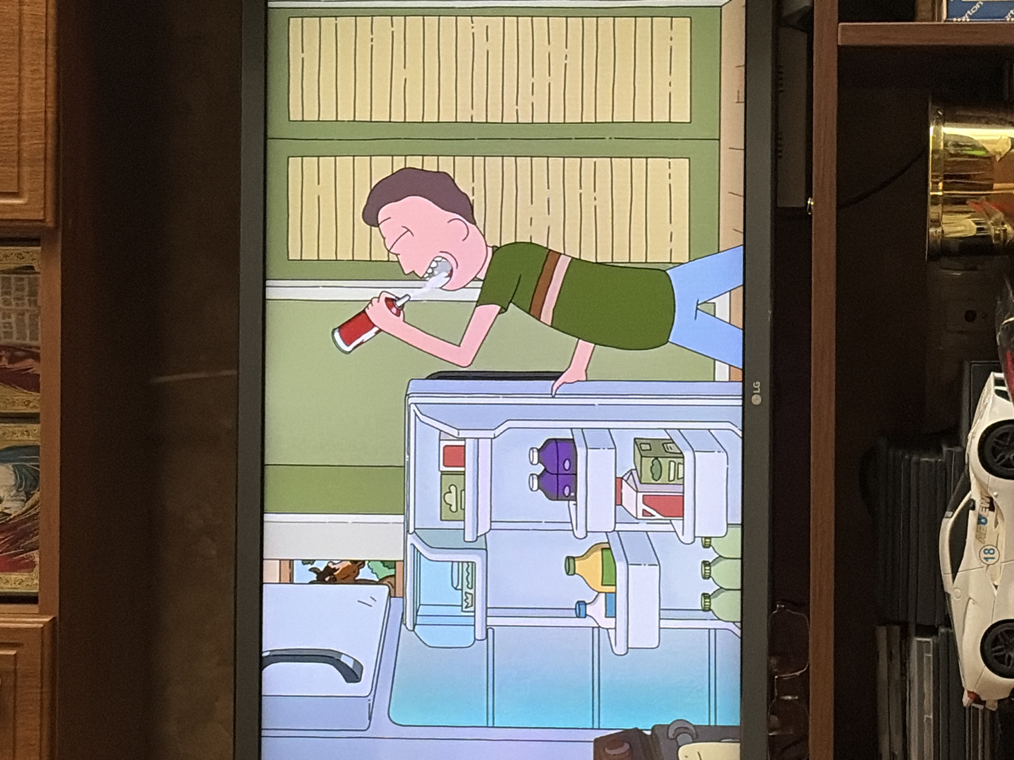 The image shows a scene from an animated television show displayed on a television screen. In the foreground, a young boy with brown hair and a green and brown striped t-shirt and blue pants is shown drinking from a red can. He is standing in front of an open refrigerator. The refrigerator is stocked with various items, including cartons of juice, bottles of milk, and other containers. Behind the refrigerator, a horse's head is visible peeking out from a doorway. The background of the scene consists of a wall with horizontal blinds. The television screen is positioned on a shelf above a lower shelf displaying various objects, including what appears to be a white model car and a brass container. The overall environment appears to be indoors, within a domestic setting.