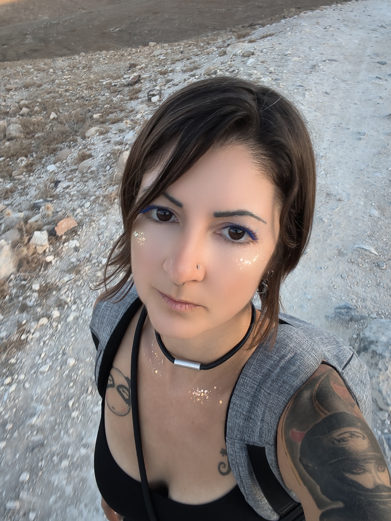 A medium shot shows a woman with brown hair and a nose ring posing for a selfie outdoors. She has blue eyeliner and glitter makeup on her face and chest. She is wearing a black tank top and a grey backpack. She has tattoos on her left shoulder and arm, and a small tattoo on her chest. The background is a dirt path with rocks and dry vegetation, suggesting a rural or undeveloped area. The lighting suggests it is late afternoon or early evening.