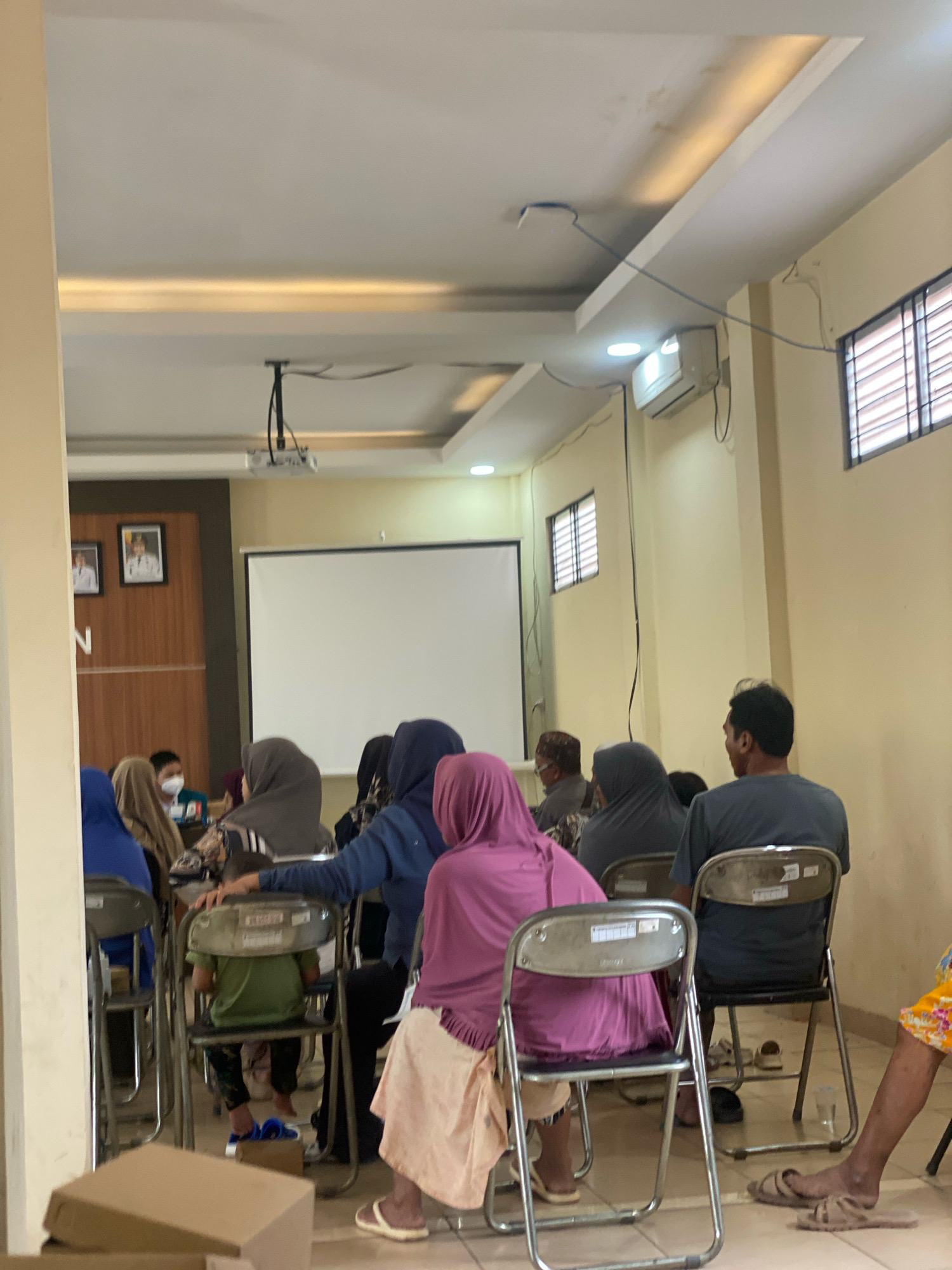 The image shows an indoor gathering of people in what appears to be a meeting room or classroom in Banjarbaru, Indonesia. There are approximately 10-15 individuals present, most of whom are seated on chairs facing a projection screen. Several people are wearing headscarves, suggesting a religious or cultural context. A projector is mounted on the ceiling, indicating a presentation or educational session is underway or has recently concluded. The room is equipped with windows and an air conditioning unit. The overall atmosphere seems to be one of focused attention or quiet observation.