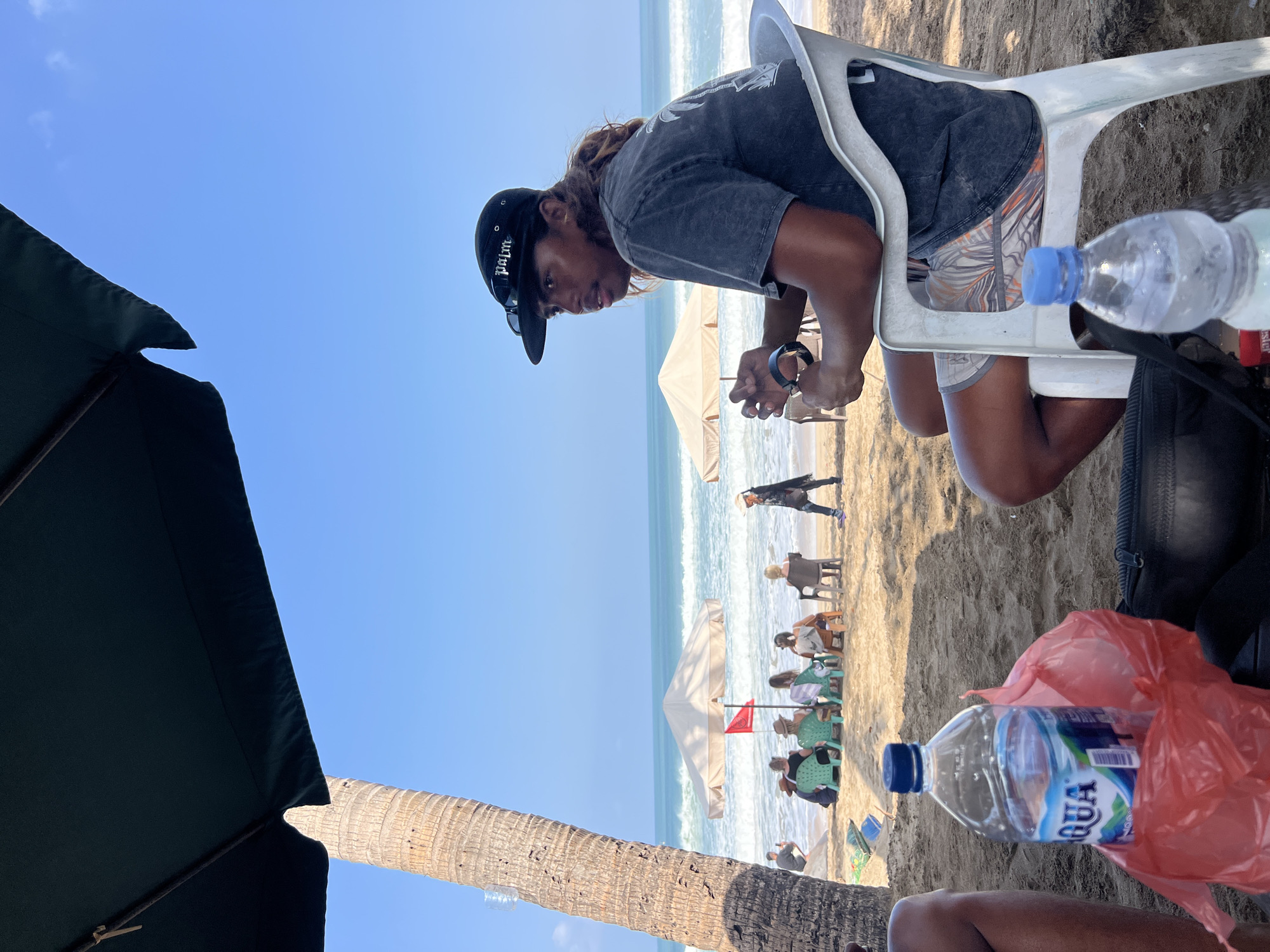 In Kuta, Indonesia, a man with long, brown hair and a black baseball cap sits in a white plastic chair on a sandy beach. He is wearing a dark t-shirt and patterned shorts, and appears to be holding something in his hands. To his right, another person's legs are visible, partially obscured. In the foreground, two plastic water bottles and a red plastic bag are placed on the sand. In the background, several people are seated in chairs under umbrellas, facing the ocean. The waves are visible, and the sky is clear and blue. A palm tree trunk is prominent on the left side of the image.