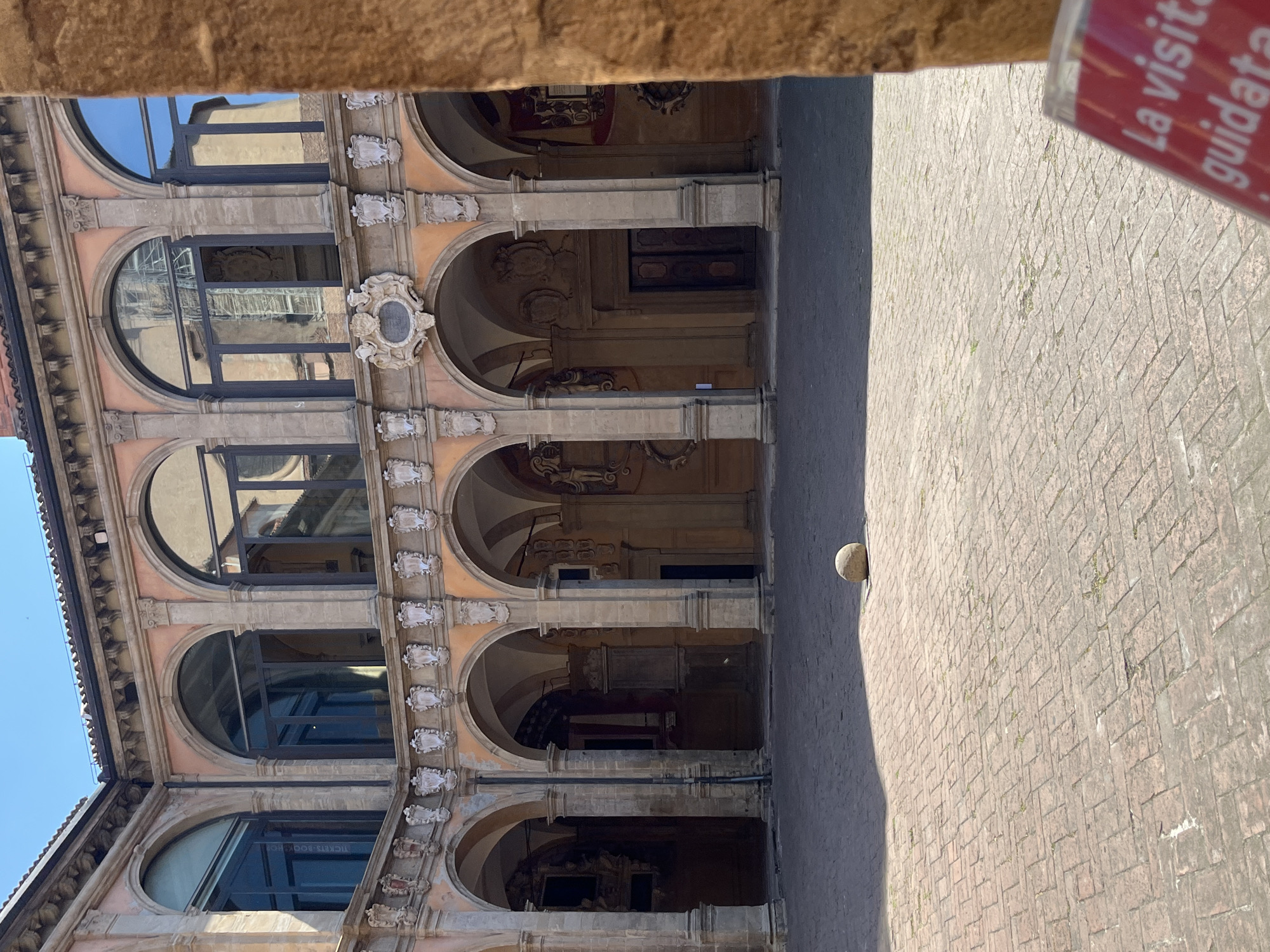 The image shows a portion of the Palazzo dell'Archiginnasio in Bologna, Italy. The building features a courtyard with arched windows and columns. The facade is adorned with decorative elements. A stone ball sits on the edge of the courtyard. Part of a sign is visible on the right.