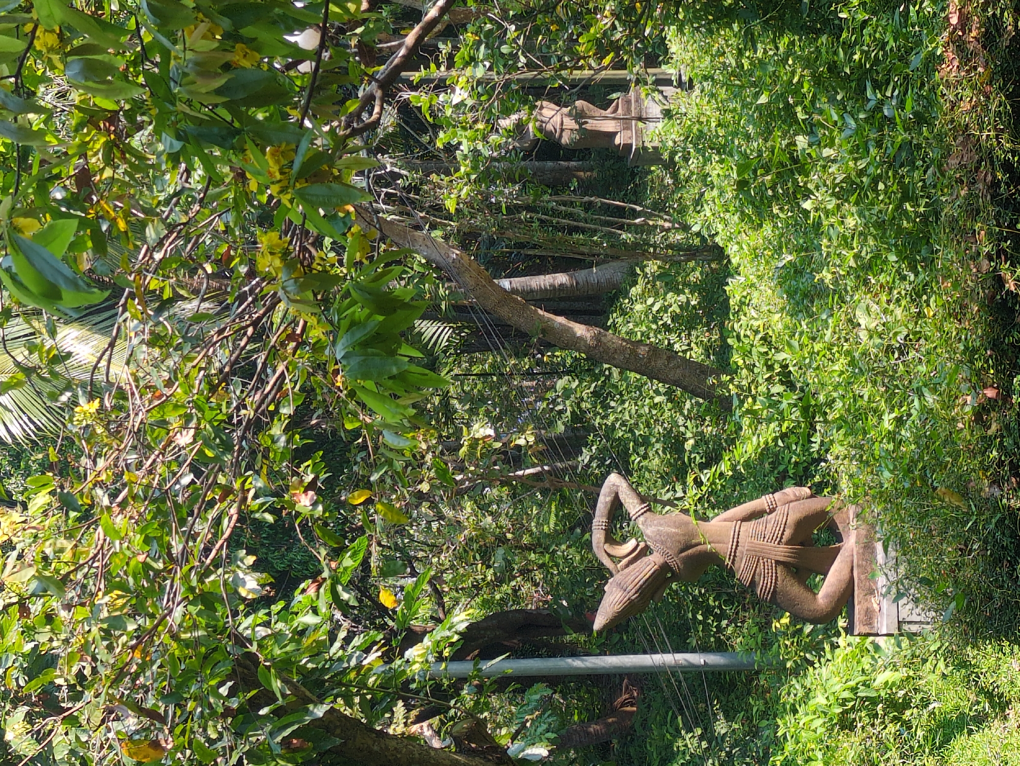 The image captures a scene in Tuy Hòa, Vietnam, focusing on a weathered stone statue of a figure in a natural setting. The statue, which has traditional clothing and ornamentation, stands among lush greenery. Other statues can be seen in the distance, creating an impression of a statue garden or area where traditional figures are displayed. The setting is outdoors, with trees and plants dominating the environment, and utility lines visible in the midst of vegetation.