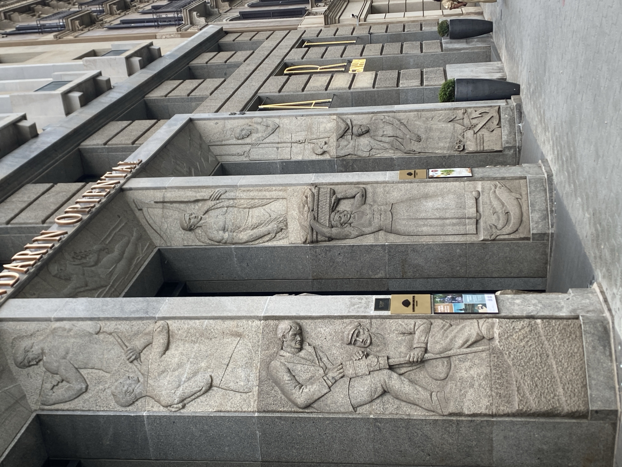 The image captures a portion of the Mandarin Oriental hotel in Barcelona, Spain. The architecture features sculpted friezes depicting figures and scenes on the facade of the building. The carvings appear to be made of light gray stone. The carvings on the frieze have a bas-relief style, portraying individuals engaged in various activities. The image is taken at a low angle, looking up at the building. There are also some balcony elements on the facade and some planters.