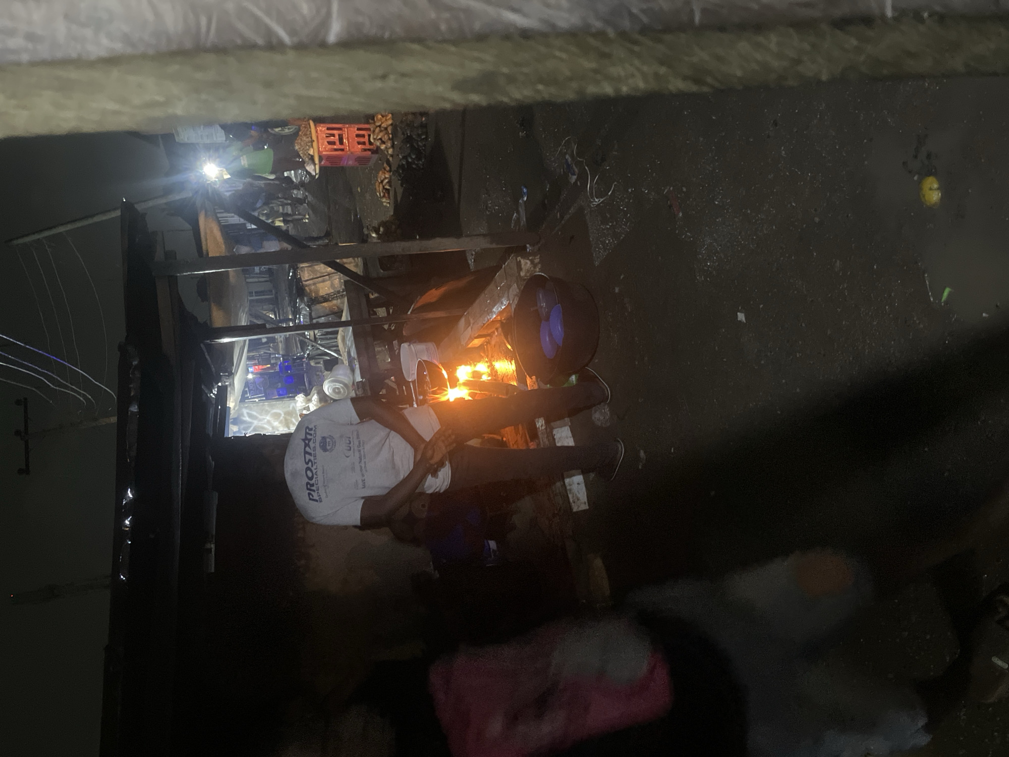 The image depicts a nighttime scene in an outdoor market area, possibly in Abuja, Nigeria. A person in the foreground is wearing a white t-shirt with "PROSTAR SPECIALTIES.COM" printed on it, along with a logo. They are standing next to a fire that is illuminating the area. Behind them, there are market stalls with various items, including red crates filled with produce, and what appear to be tubers or root vegetables scattered on the ground. There are other people visible in the background, some illuminated by lights from the stalls and the fire. The ground appears wet and possibly dirty, with some debris and a yellow object visible on the ground in the lower right portion of the image. A partially visible person on the left is wearing a pink and dark-colored garment. The overall impression is that of a busy, dimly lit market environment at night.
