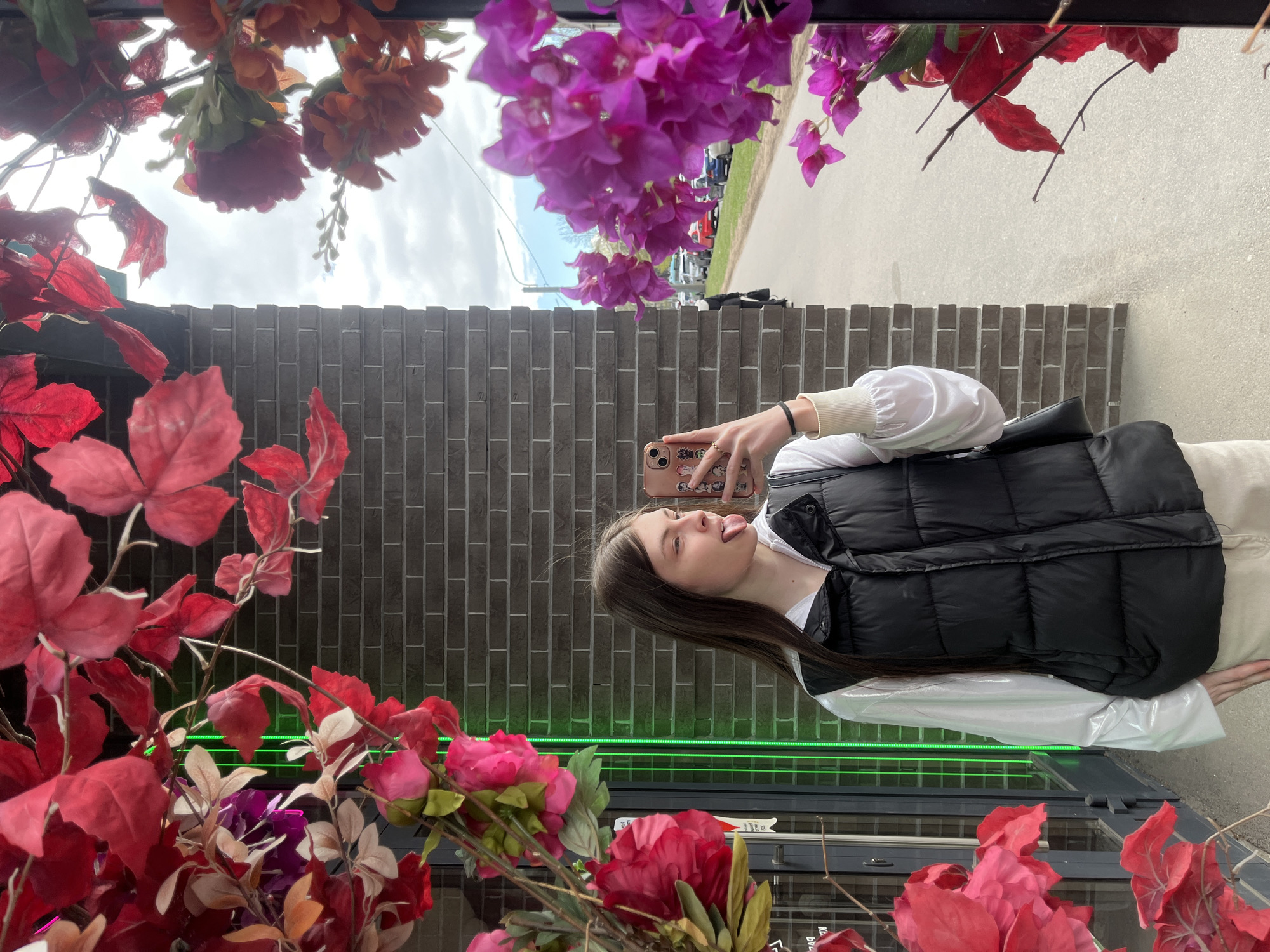 A young woman is captured in a playful mirror selfie, extending her tongue slightly. She holds a smartphone with a decorated case in her right hand, which has a black bracelet and a gold ring. She is dressed in a white long-sleeved top with cuffed sleeves and a black puffer vest. Her long brown hair falls down her back.
The setting appears to be a street-facing reflective surface, such as a large window or mirror, heavily decorated with artificial red, purple, and orange flowers and foliage, which frame the image. Behind the woman, a textured gray brick wall is visible.
Reflected in the upper portion of the mirror are an overcast sky, a road, and parked cars, suggesting an outdoor urban environment during the daytime. A prominent horizontal strip of neon green light runs across the middle of the brick wall area. Partially visible Cyrillic text, "БУЛ" and "ДУ", is reflected in the bottom right corner, possibly from a sign or window sticker. The scene suggests a casual moment in Saint Petersburg, Russia.