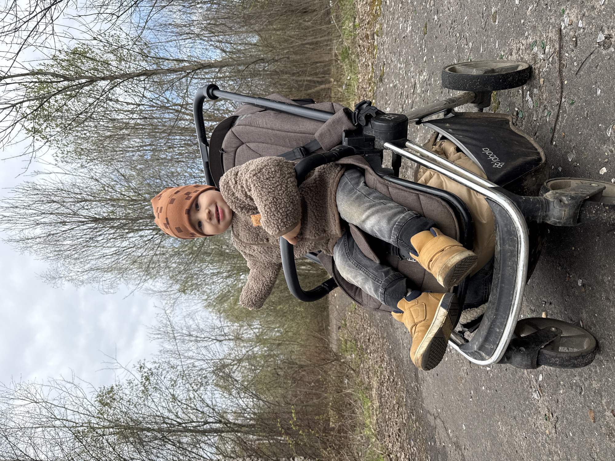 A toddler, wearing a brown patterned hat, a tan sherpa jacket, dark gray jeans, and tan boots, sits in a brown and silver stroller on a cracked asphalt path. The stroller is oriented towards the right side of the frame. The child is facing the camera with a small snack in their hand. The path is flanked by trees without leaves, creating a blurred background of branches. The sky is overcast. The word "Bobola" is visible on the side of the stroller. The exact location cannot be determined from the given information.