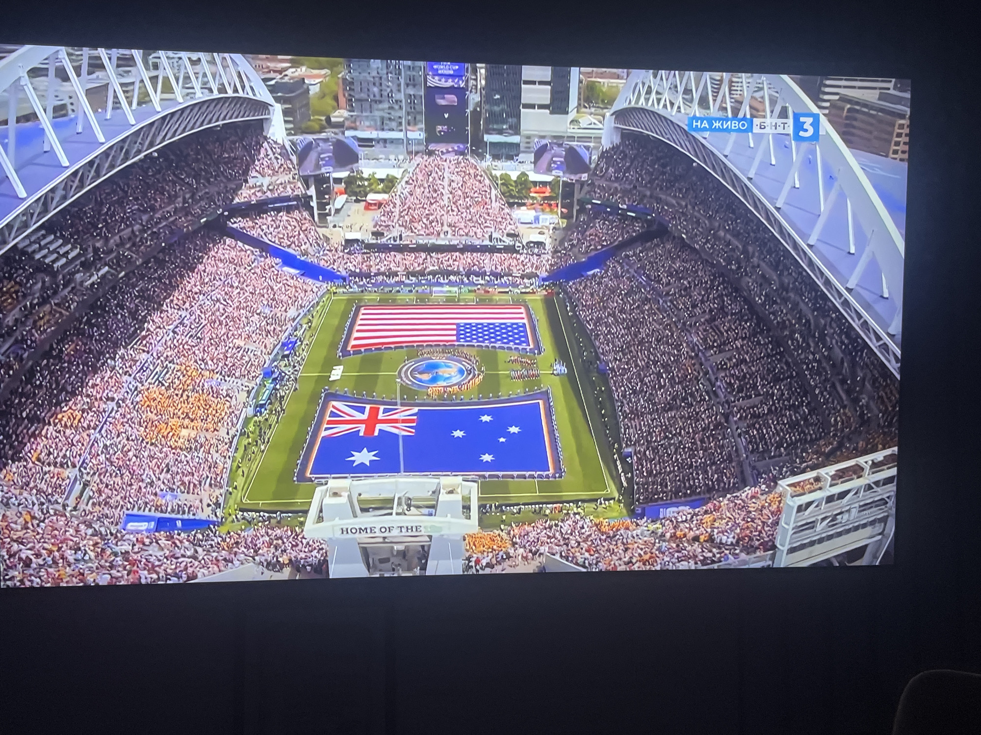 The image shows an aerial view of a packed stadium during a sporting event. Two large flags are displayed on the field: the United States flag and the Australian flag. The stadium is filled with spectators, and a large screen is visible in the background displaying "WORLD CUP". Text overlay on the top right of the screen reads "НА ЖИВО Б Н Т 3", which indicates a live broadcast on BNT 3, a Bulgarian national television channel. The stadium architecture suggests it is a modern sports venue. While the image depicts a sporting event with international flags, there is no information within the image to suggest the location is in the city of Varna, Bulgaria. The presence of the Bulgarian television broadcast information is the only indicator of a potential connection to Bulgaria.