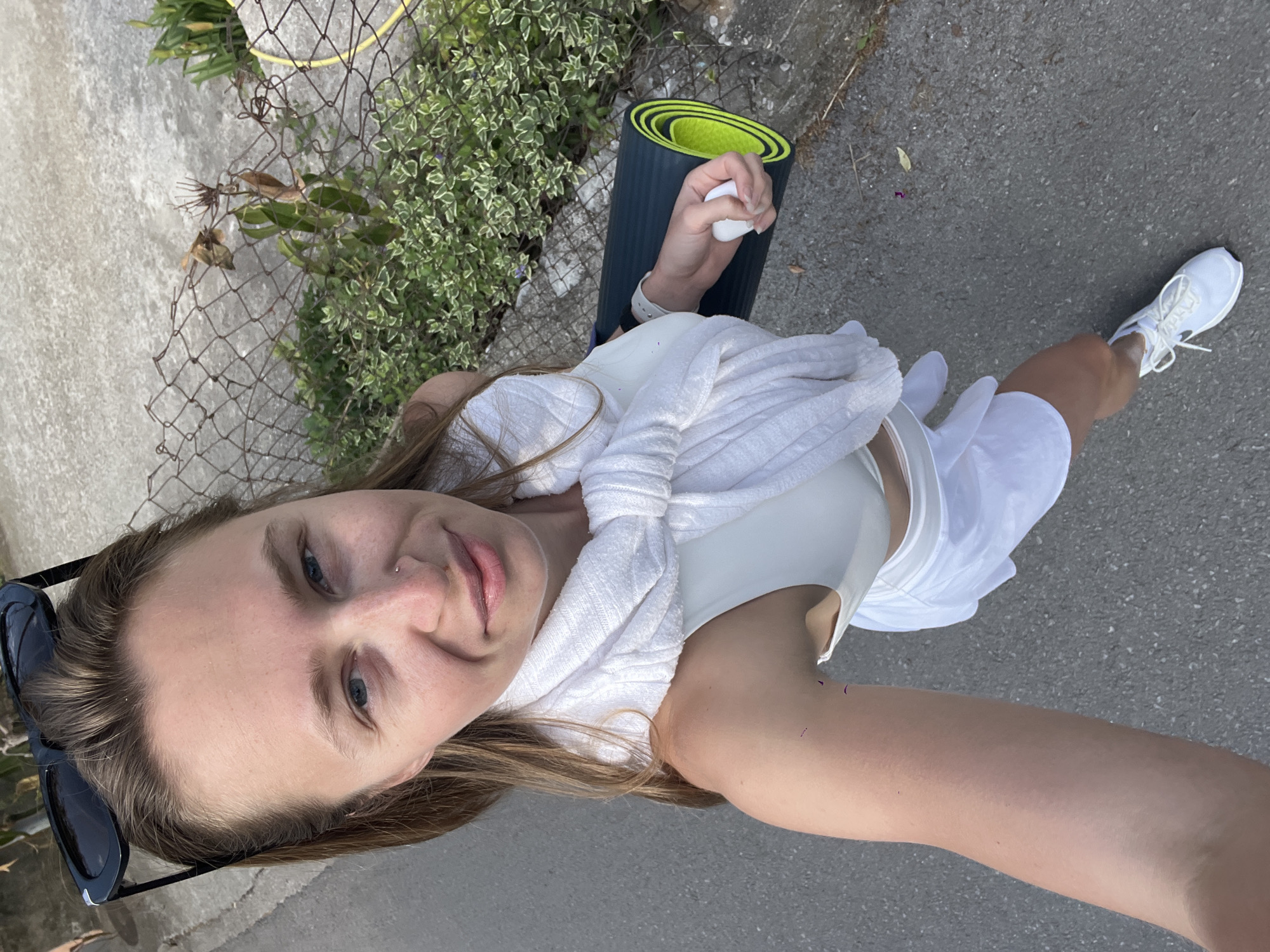 This bright, high-angle selfie shows a young woman outdoors, with the image rotated 90 degrees clockwise. The woman, who is the central subject, has light brown hair and blue eyes, and looks directly at the camera with a gentle smile. She is dressed in white athletic wear, including a sports bra or tank top, shorts, and a textured white sweater or shawl tied around her shoulders. Black sunglasses are pushed up onto her head, and a small nose piercing is visible on her left nostril. On her left wrist, she wears a white watch, and white sneakers are on her feet.
In her right hand, she holds a rolled-up dark blue yoga mat with a bright green interior, along with a small white object, possibly an earbud case. The setting is an outdoor paved area, likely a path or sidewalk, with light grey asphalt or concrete underfoot. To her left, there is a chain-link fence, some parts appearing rusted, with various green plants and variegated foliage growing through and behind it. Beyond the fence, a light-colored concrete wall or surface is visible.
The clear, soft lighting suggests it is daytime, possibly morning or late afternoon, indicating pleasant weather. Her attire and the yoga mat imply she is either heading to or returning from a fitness activity, such as yoga, in Bečići, Montenegro. Her expression conveys a relaxed and happy disposition. There is no visible text in the image.