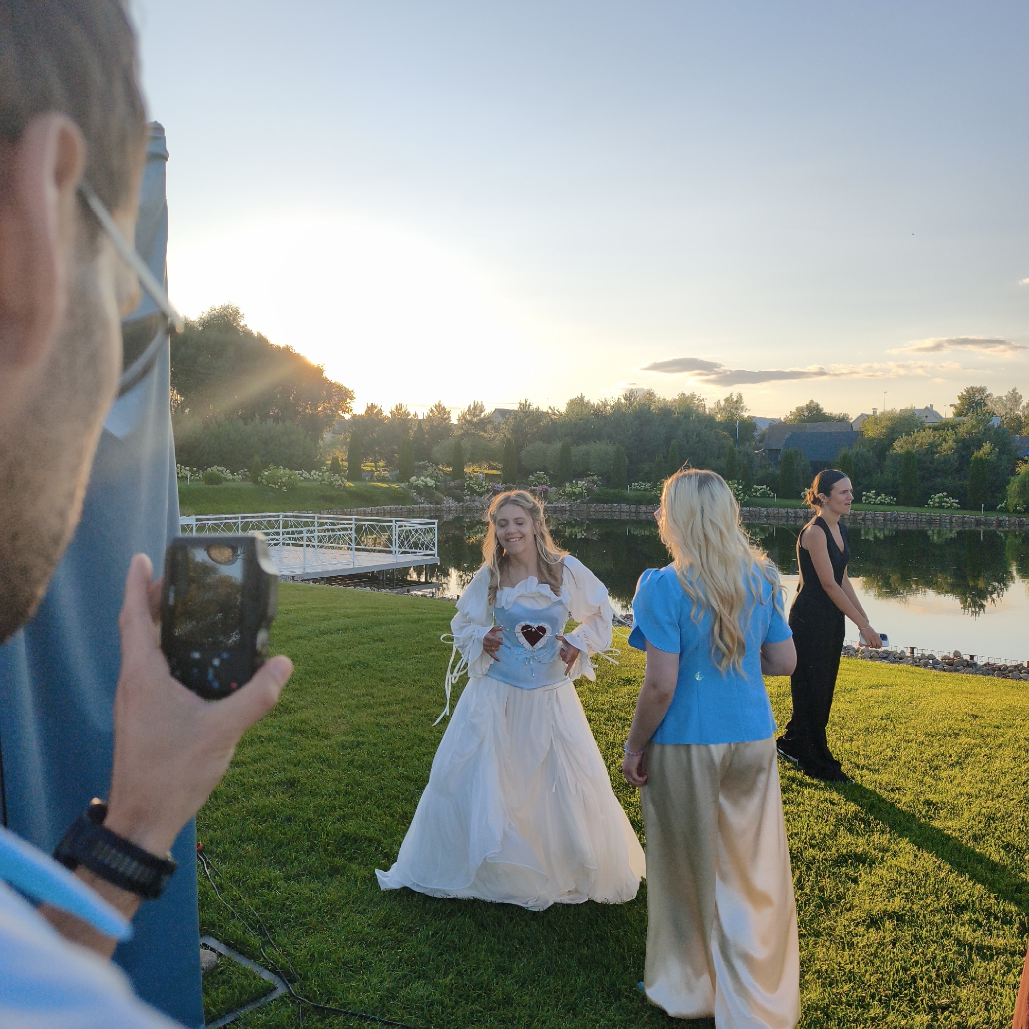 In Sapotskinski Village Council, Belarus, a group of people are gathered outdoors near a body of water. In the foreground, a person's arm and hand are visible, holding a dark-colored mobile phone, seemingly taking a photograph. The phone's screen reflects trees and the sky. To the left, a portion of a blue fabric structure is also visible.
Further into the scene, a woman in a white, flowing dress with a blue corset adorned with a heart is standing on a green lawn. She is looking downwards with a slight smile. Beside her, another woman in a bright blue top and cream-colored pants faces away from the camera. In the background, a woman in a black dress is standing.
The environment features a calm body of water with a white railing structure extending over it, possibly a pier or walkway. Trees and bushes line the edge of the water, with more trees and some houses visible in the distance. The sky is a clear blue with a gentle sunset casting warm light across the scene. The overall atmosphere suggests a casual gathering or an event taking place outdoors in the late afternoon or early evening.