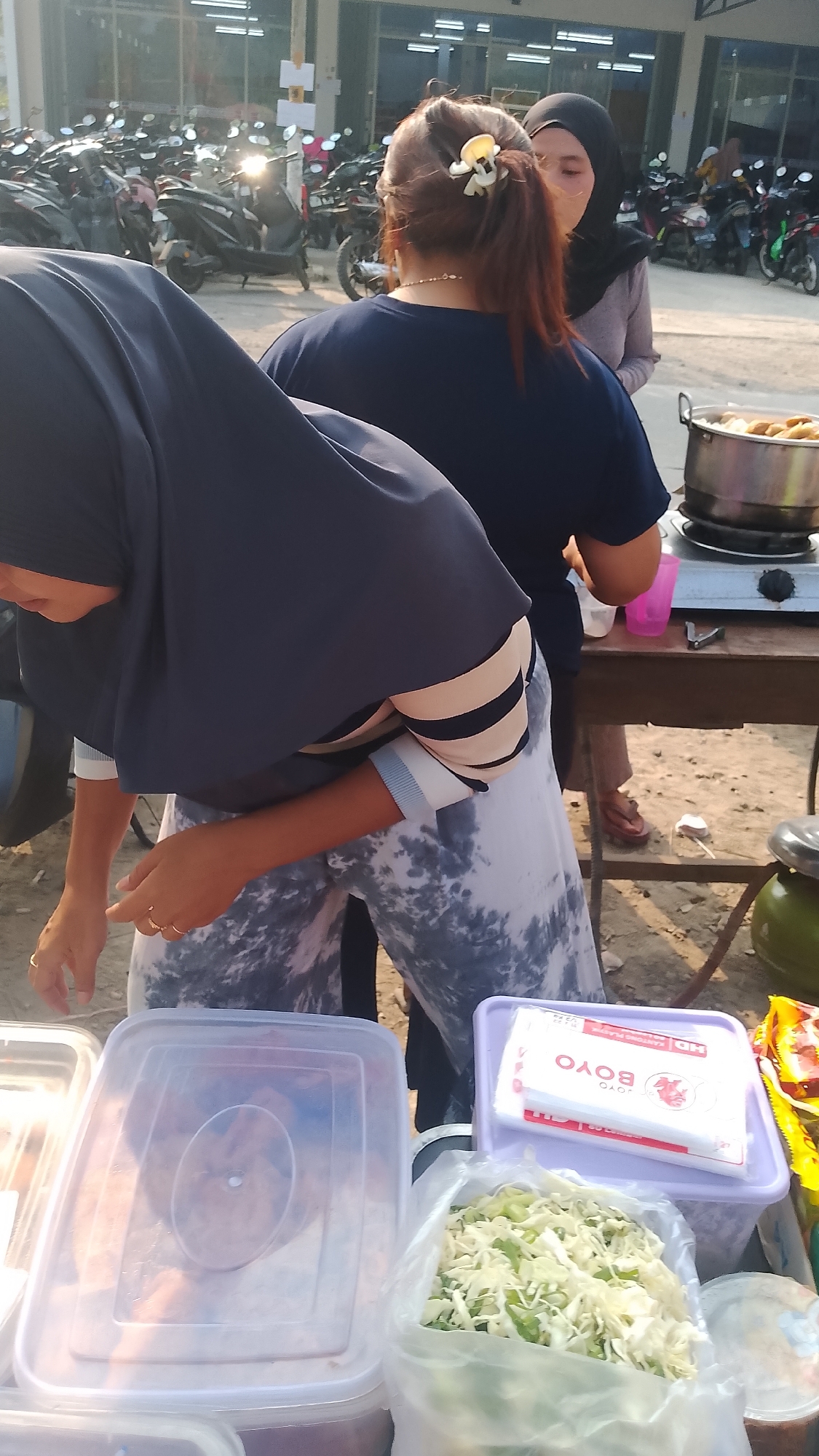 The image captures an outdoor scene at what appears to be a street food stall in Karanglangu, Indonesia. In the foreground, a person wearing a dark blue hijab and a striped long-sleeved shirt is bending over, reaching towards a clear plastic container filled with chopped green vegetables. Next to it, a purple container holds a stack of white paper napkins with red lettering. To the left, another large clear plastic container is visible, partially obscuring a person wearing a dark blue head covering.
In the mid-ground, a table is set up with a gas stove and a pot containing food, possibly meatballs or dumplings. A pink cup and some utensils are also on the table. Another person, wearing a grey long-sleeved shirt and a black hijab, is standing behind the table, looking towards the front.
The background reveals a multitude of parked motorcycles, suggesting this location might be a hub of activity or a marketplace. The overall environment is somewhat dusty and unpaved, consistent with an outdoor market or roadside setting in Indonesia. The sunlight indicates it is daytime.