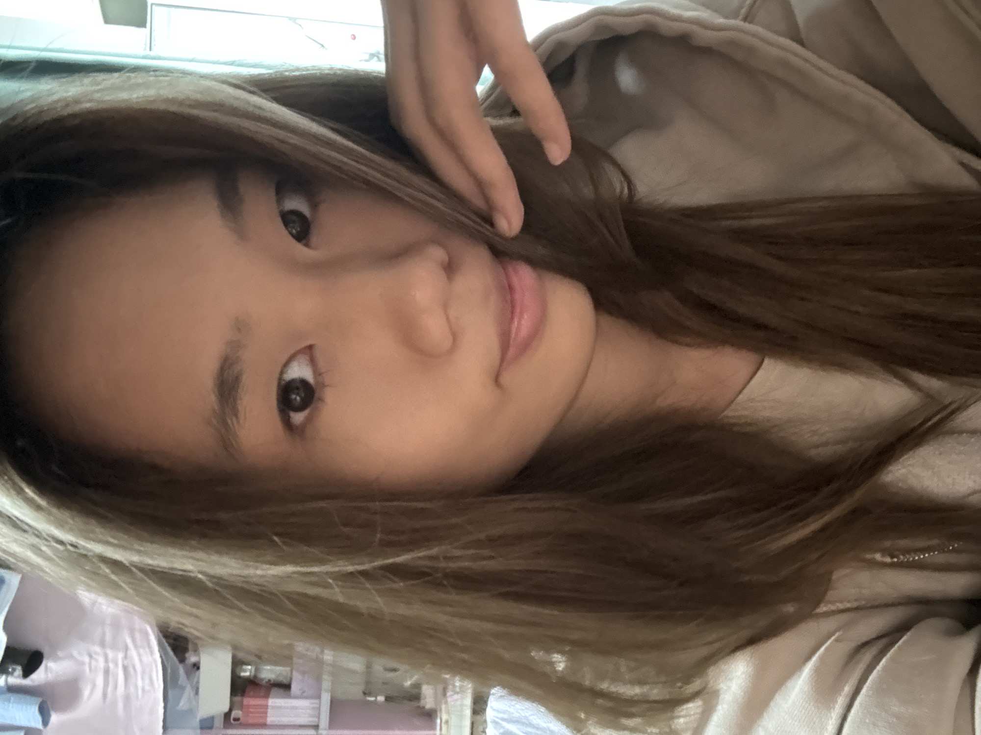 A close-up portrait shows a young Asian woman indoors, possibly in a residential space based on the glimpse of furniture and household items in the background. Her hair is light brown, and she has one hand near her face. She appears to be wearing a light-colored jacket or sweater. The photo is taken at an angle, not straight-on. The location in the city of Nan'an District, China, cannot be confirmed based on visual information in this image. There is no definitive evidence of location.