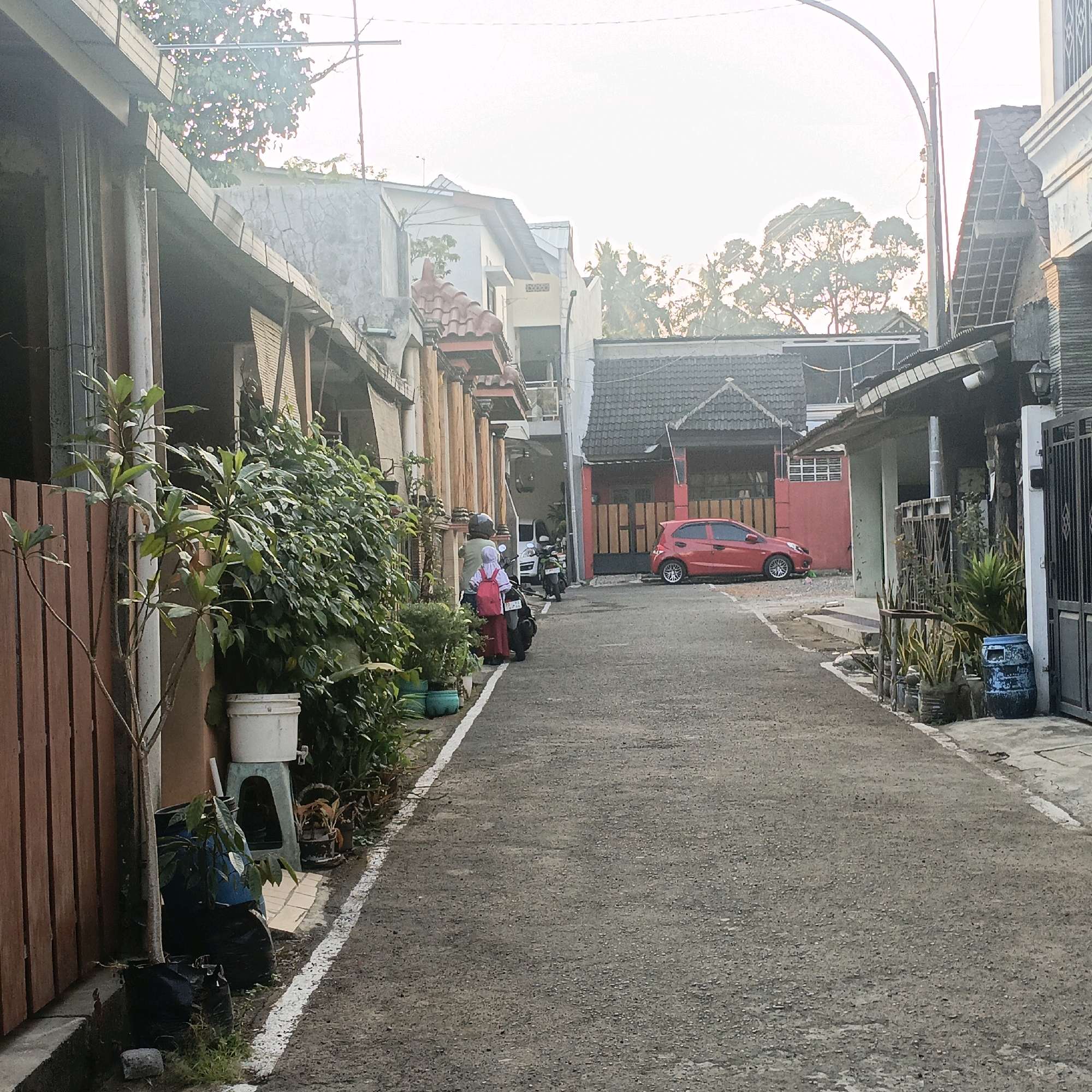 The image depicts a narrow asphalt road in Salatiga, Indonesia, flanked by residential buildings and dense vegetation. A red car is parked on the right side of the road. On the left, a person on a scooter wears a headscarf and a backpack. Several potted plants and a blue barrel are visible along the roadside. The scene suggests a typical urban residential environment in Indonesia.