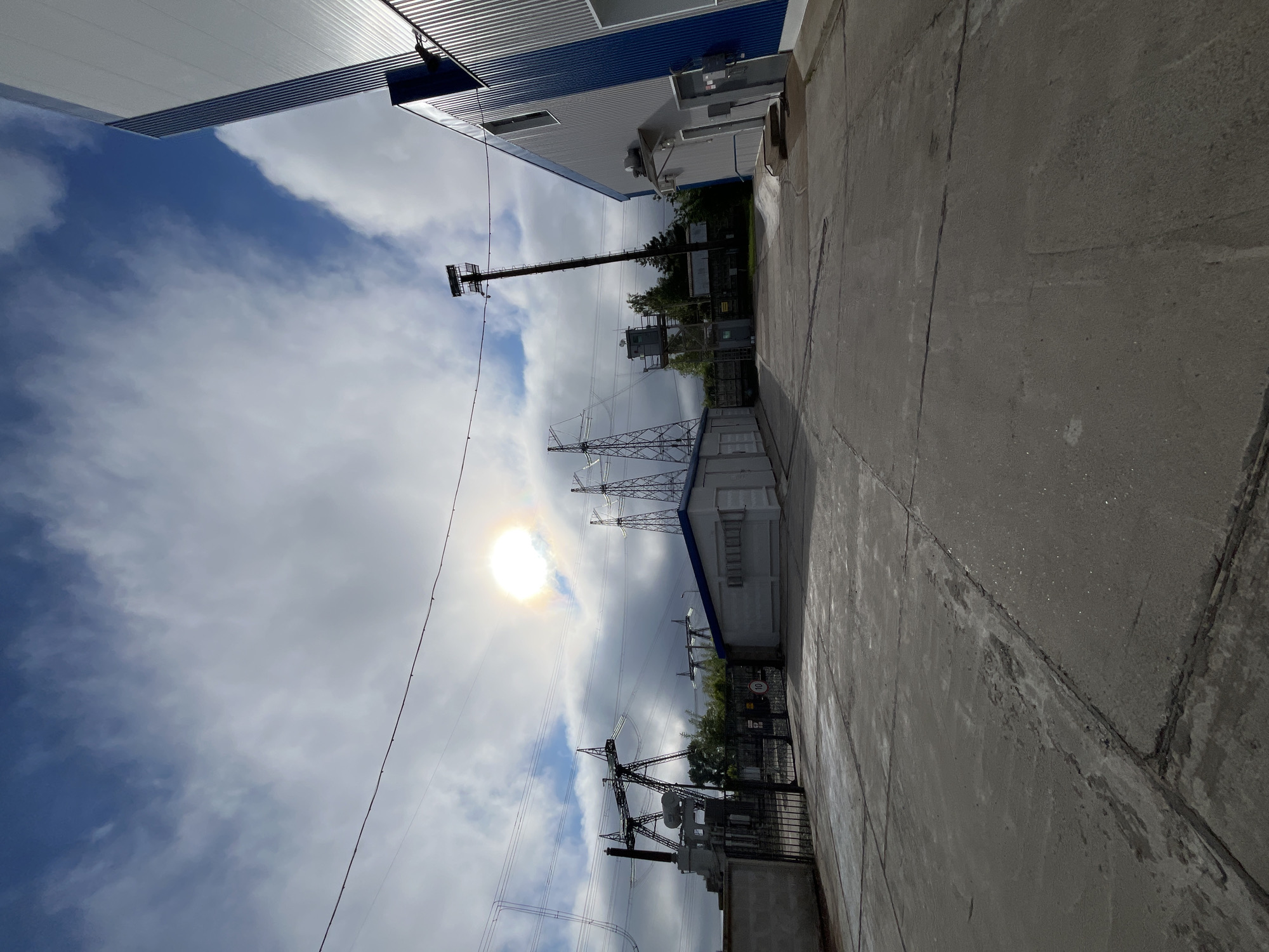 The image depicts an outdoor industrial area under a cloudy sky with the sun partially obscured by clouds, creating a bright halo effect. In the foreground, a concrete surface is visible, marked with cracks and some areas of discoloration. To the left, a metal fence encloses a section of the area, which includes a substation with transformers and high-voltage power lines supported by metal towers. A small, white building with a blue roof is situated behind the fence. Further to the right, a large, modern building with corrugated metal siding, predominantly gray with blue accents, runs along the edge of the frame. A tall, dark utility pole with a square platform at the top stands between the substation and the modern building. In the background, trees and other industrial structures are present. The scene appears to be located within an industrial zone, possibly a power substation facility, in the city of Chaikovsky, Russia. There are no people visible in the image.