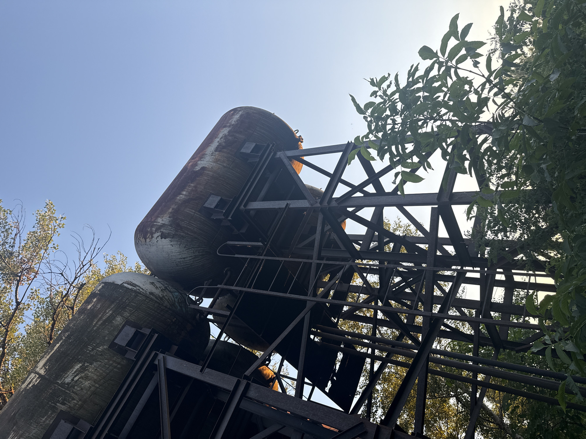 The photograph, taken from a low angle looking upwards, depicts a rusty, elevated water tank structure in Obukhivka, Ukraine. Two large, cylindrical tanks, heavily weathered and stained, are supported by a complex framework of dark metal beams and supports. A ladder extends up the side of one of the tanks. The structure is partially obscured by green foliage and trees, with dappled sunlight filtering through the leaves. The sky above is a clear, pale blue. The overall impression is one of disuse and decay, with the natural elements beginning to reclaim the industrial infrastructure.