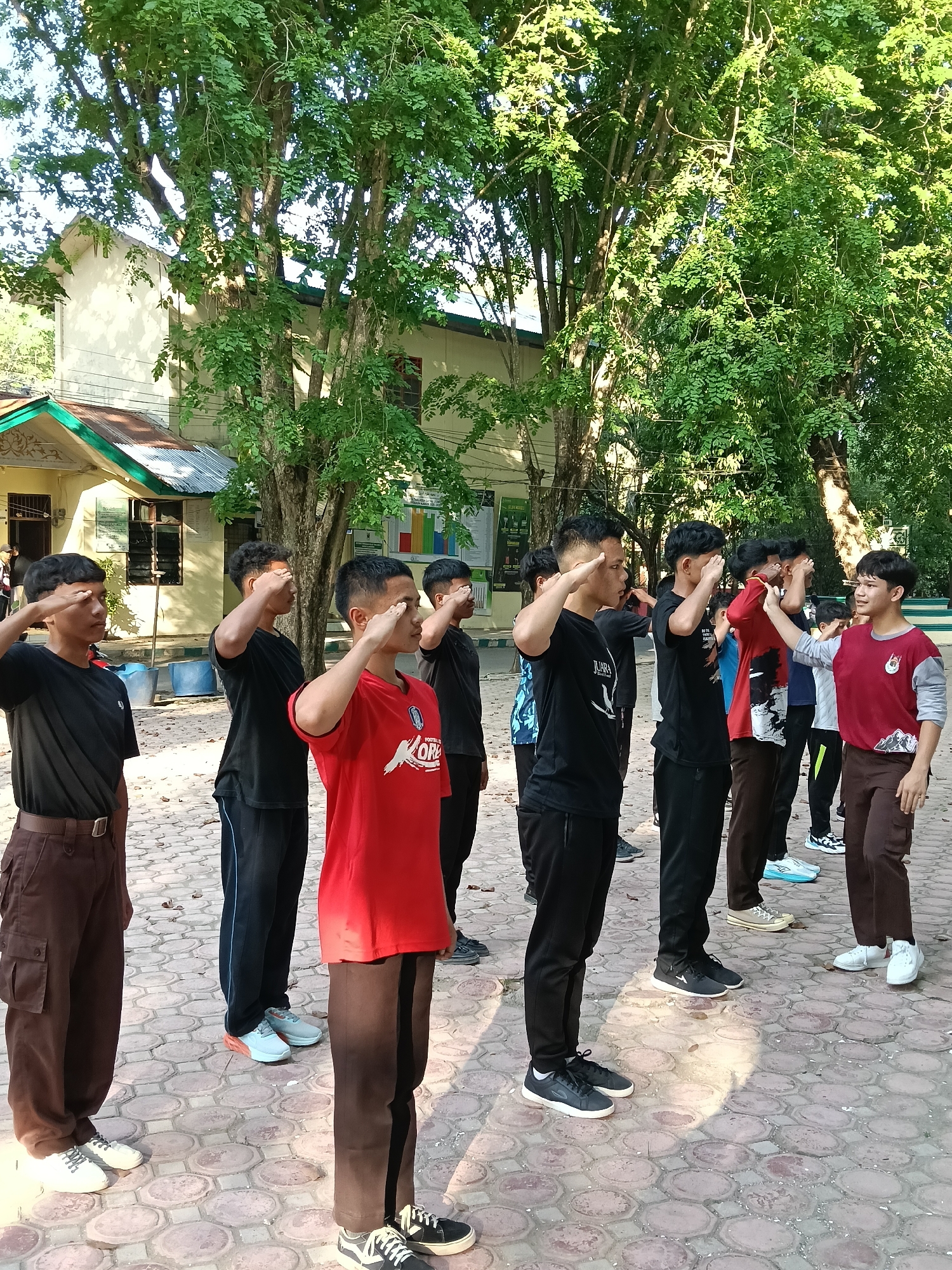A group of approximately ten young men stand in a line in an outdoor area. They are performing a salute, with their right hands raised to their foreheads. The young men are wearing a variety of casual clothing, including t-shirts, pants, and sneakers. The man closest to the viewer is wearing a black t-shirt, brown cargo pants, and white sneakers.
The setting appears to be a paved courtyard or open space, with trees visible in the background. A single-story building with a pitched roof can be seen behind the trees. The building appears to be in Lhokseumawe, Indonesia. The area is well-lit, suggesting it is daytime. The scene conveys a sense of order and discipline.