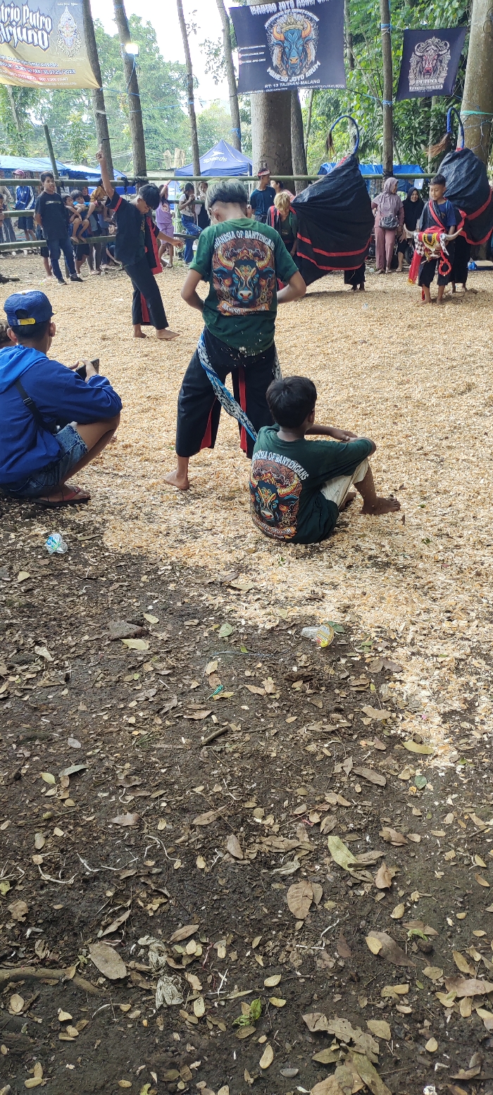 The image shows an outdoor scene in a park, likely in Kecamatan Lawang, Indonesia, with the given coordinates. There are a group of people in the scene, some wearing traditional Indonesian clothing. Two people in the foreground are wearing green shirts with a logo that says "ANNISA OF BANTENGANS". The people are standing on a dirt path with brown leaves scattered about. There is a black tent in the background, and a sign that says "BANDORO JAYA AUTO". The scene is likely during a public event or gathering. There are some items of litter on the ground including a plastic bottle and plastic bag. There are trees in the background and a metal ring in the upper middle of the picture.