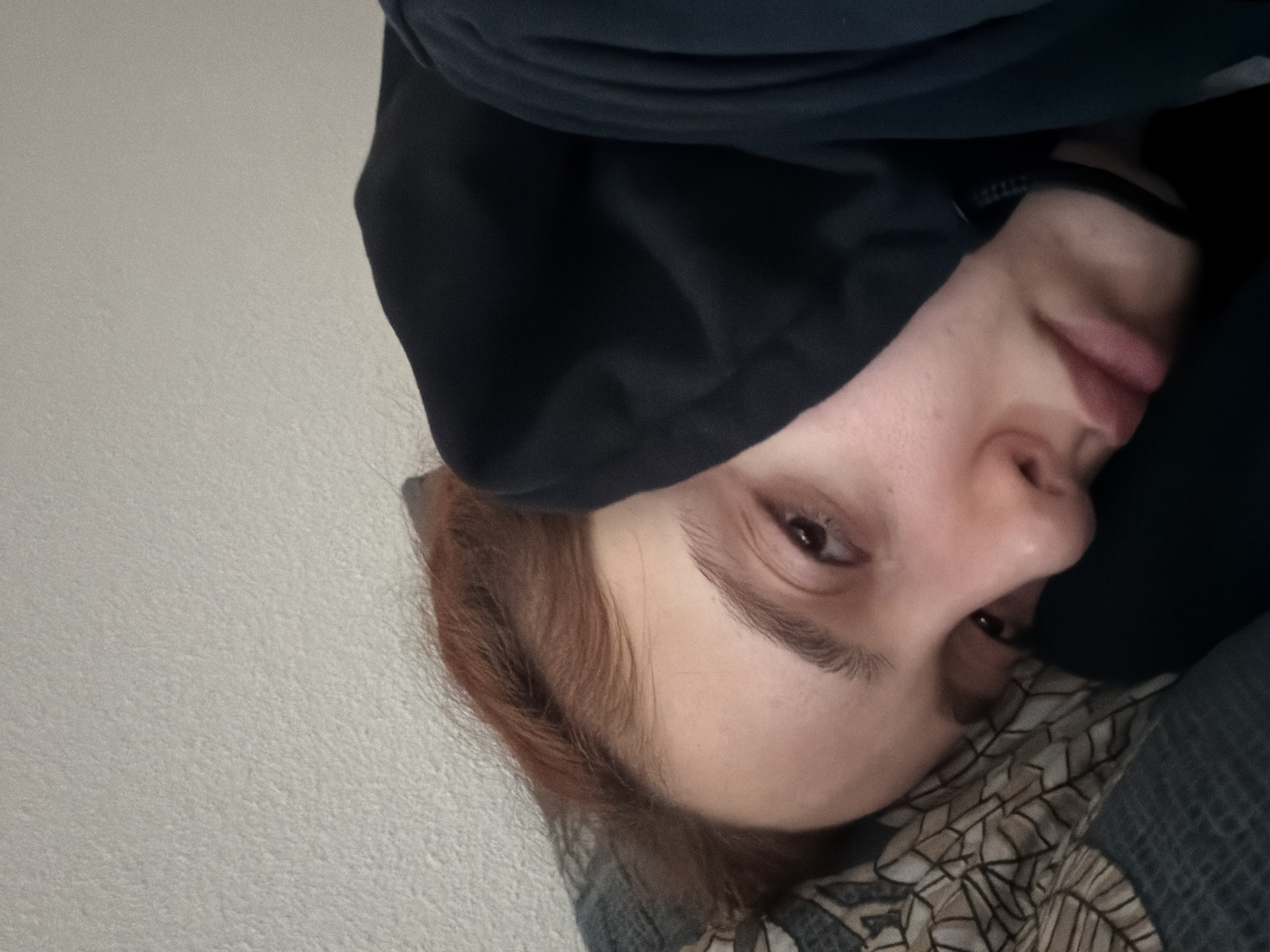The image depicts a close-up, angled view of an individual, appearing female, lying down indoors. The scene is located in the city of Strasbourg, France. The subject's head rests on a pillow covered with a patterned fabric featuring dark geometric shapes on a light background. She has reddish-brown hair, visible on the upper left, and light skin. She is wearing a dark-colored, likely black, hooded garment, which is partially pulled up over her head. A black strap or choker is visible around her neck, and a lighter-colored undergarment is partially visible at the collar area beneath the hoodie. Her face is oriented towards the camera, with her eyes open and a neutral expression. The background consists of a light-colored, textured wall, out of focus. The lighting is ambient and even. No other individuals or identifying objects are present.