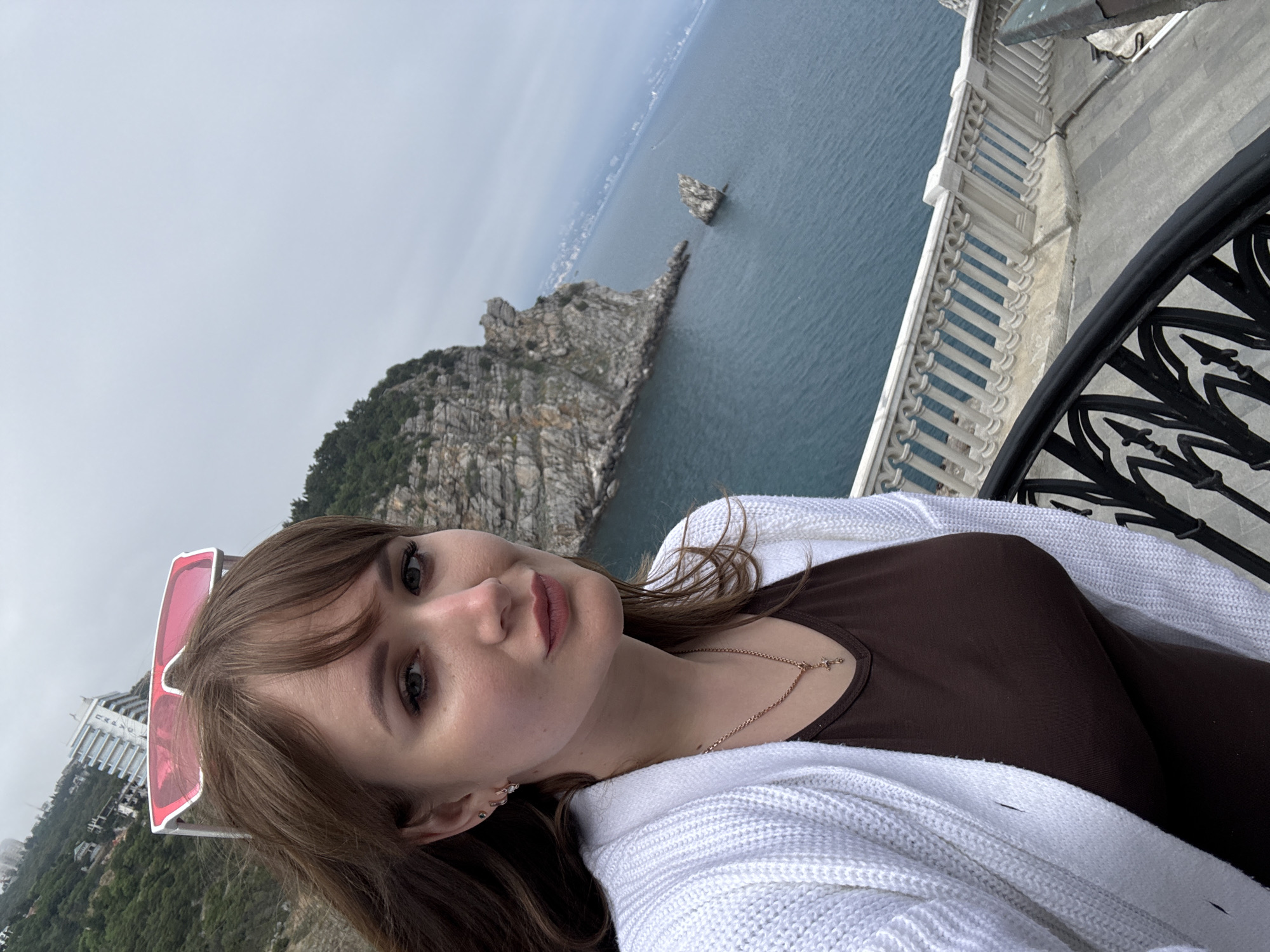 A woman stands on a stone balcony with a wrought iron railing, posing for a selfie. The background features a coastal landscape in Гаспринский поселковый совет, Ukraine, with rocky cliffs, a body of water, and a distant building. The woman is wearing a brown top and a white cardigan. She has brown hair with bangs and is wearing pink sunglasses pushed up on her head. A gold necklace is visible around her neck. The sky is overcast.