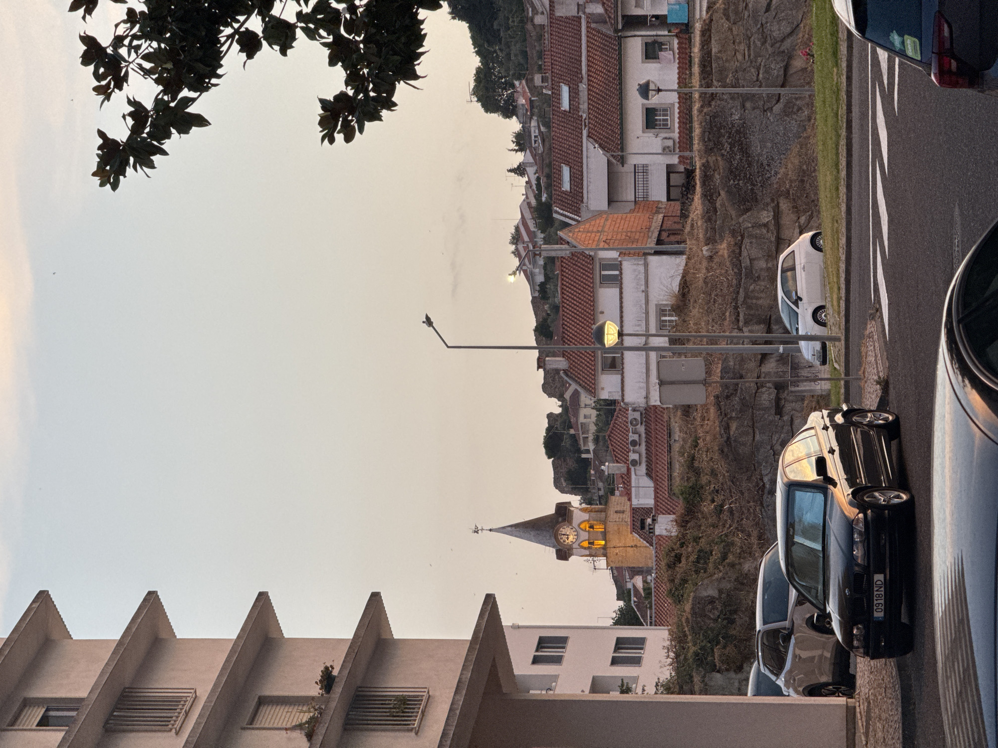 The photograph captures a street scene in Castelo Branco, Portugal, during the golden hour. In the foreground, a black sedan with license plate "0918 ND" is parked on a paved surface. To its left, a white SUV is partially visible. Behind the cars, a steep, rocky embankment is covered with dry vegetation.
Dominating the mid-ground is a cluster of buildings with red-tiled roofs, characteristic of Portuguese architecture. One building features exposed brickwork. Further back, a church tower with a clock face is illuminated from within, its spire reaching towards the sky. The background reveals a hillside dotted with more houses, their roofs also red-tiled, and lush green trees. The sky above is a soft, pale orange, indicating the setting sun. A street lamp stands prominently in the mid-ground, its light illuminating the foreground. A large tree with dark green leaves is silhouetted in the upper right corner of the frame. There are no people visible in the image.