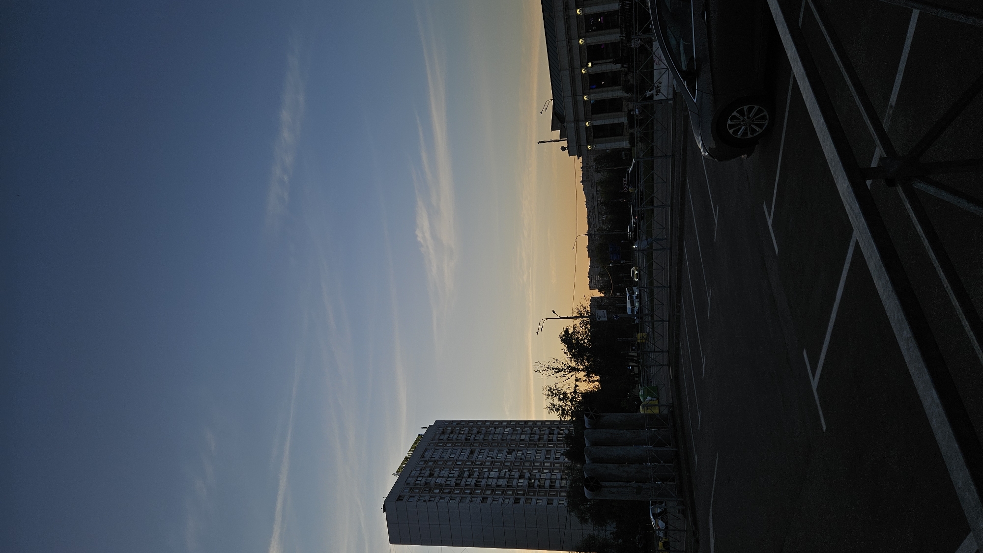 The image shows an outdoor scene at dusk in an urban area. A tall, rectangular building with many windows occupies the left side of the frame. The building has a sign on top in a foreign language. The sky is a gradient of blue to orange, indicating sunset or sunrise. To the right of the building, there are trees and streetlights. Adjacent to the trees, there is a parking lot with marked spaces. A car is parked in one of the spaces, partially visible in the frame. The scene is viewed from an elevated perspective, with a fence visible in the foreground. The location is identified as the city of Unknown, Unknown.