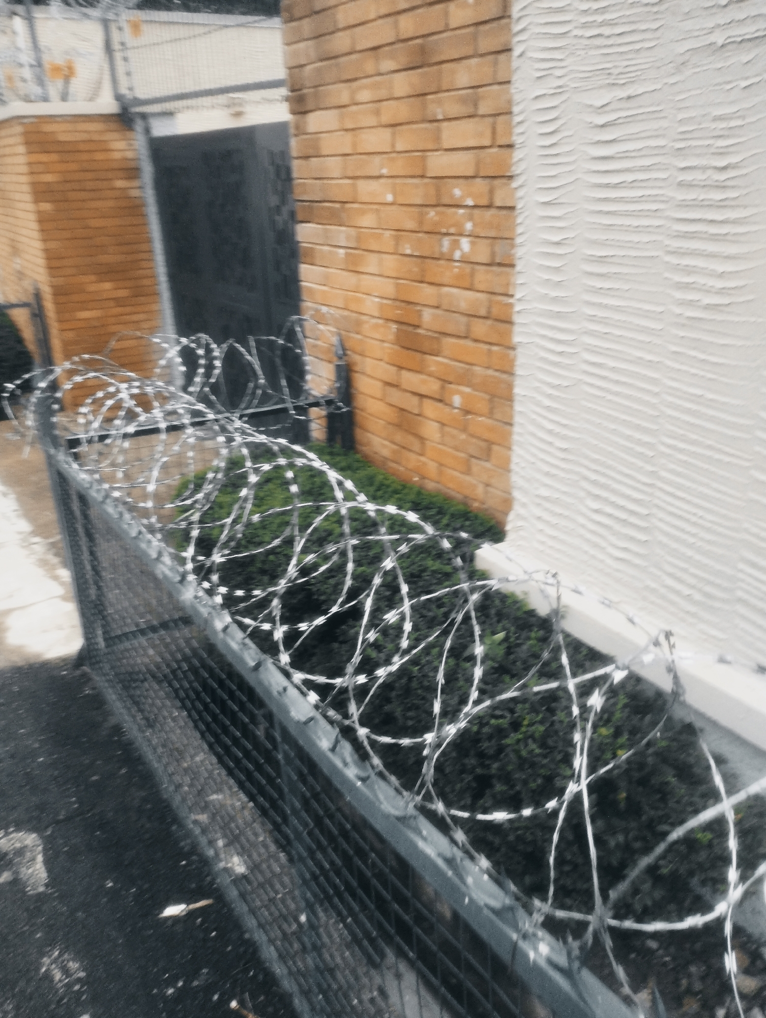 The image depicts a security fence topped with concertina wire, situated in front of a building constructed with orange bricks and white stucco. The fence is made of metal mesh and metal posts. Behind the fence, a dense green hedge is visible, and further back, a dark, recessed doorway is present within the brick wall. The ground in front of the fence appears to be asphalt or concrete. The scene is likely an outdoor area, possibly a perimeter or entrance to a property. There is no indication of the city of Jos, Nigeria, in the image.
