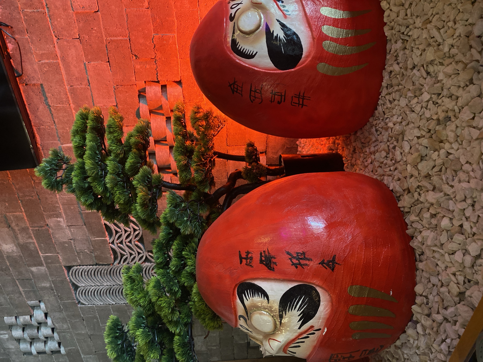 The image shows two large, red Daruma dolls with painted faces and Japanese script, stacked vertically against a backdrop of a brick wall on the left and white gravel on the right. A small, stylized evergreen tree is positioned to the left of the lower doll. The scene is illuminated by a reddish-orange light source. The location is indicated to be in Krasnoyarsk, Russia, based on additional information not directly visible in the image.