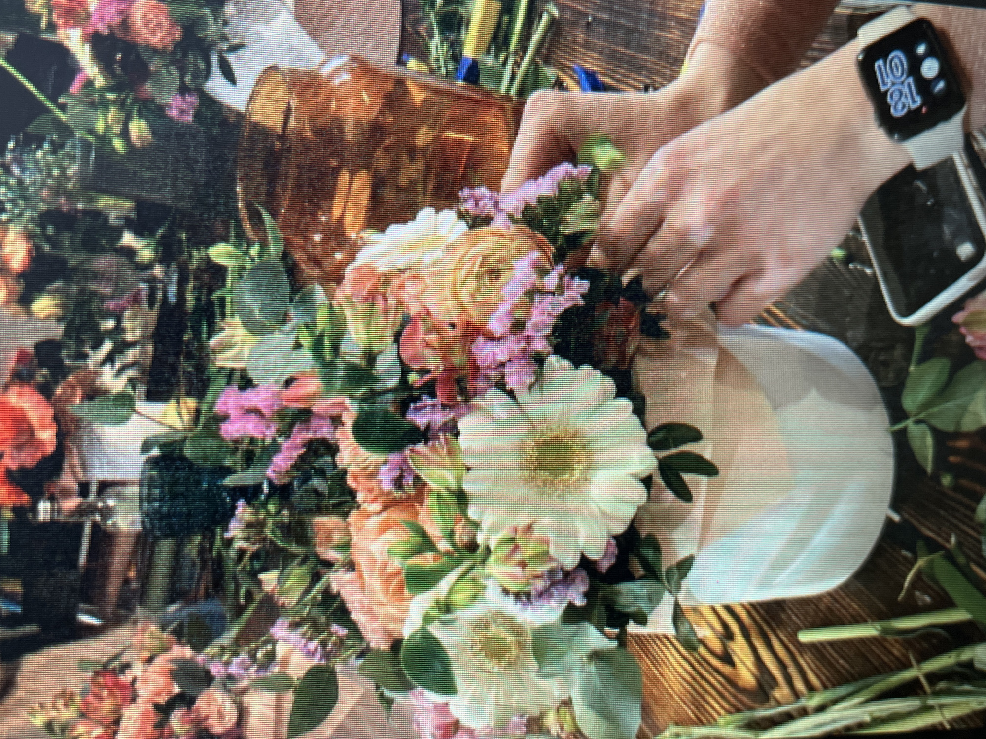 In the city of Sejahtera, Indonesia, a close-up shot captures a florist arranging a vibrant bouquet of peach, white, and pink flowers in a white cylindrical vase. The arrangement includes large white gerbera daisies and peachy ranunculus, accented by delicate pink spray flowers and lush green foliage. Two hands are visible, actively tucking in stems to complete the bouquet. Behind the arrangement, a large, amber-colored glass vase and various floral arrangement tools are partially visible. The setting appears to be a workshop or studio, indicated by the wooden tabletop surface. A smartwatch is worn on the right wrist of one of the hands, displaying the time as 10:09.
