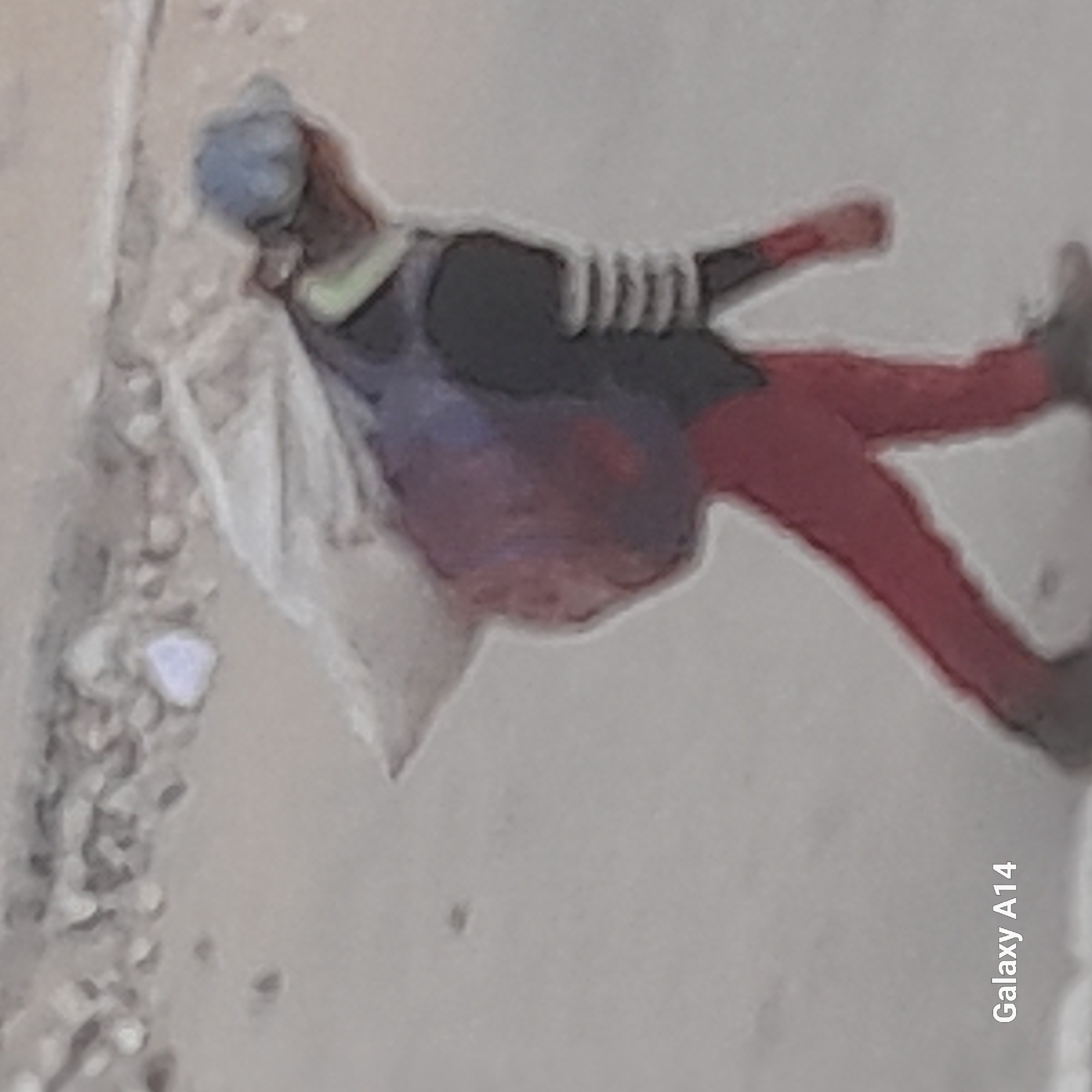 An image shows a person walking in what appears to be an outdoor, possibly urban, environment. The person is carrying a large, bulky bag or bundle on their back, which is mostly covered by a white fabric. They are wearing a light blue cap, a dark jacket with lighter striped sleeves, and red trousers. The ground appears to be sandy or dusty. The "Galaxy A14" watermark is present in the bottom left corner, indicating it is a mobile phone image. The scene suggests a person engaged in transport or carrying goods.