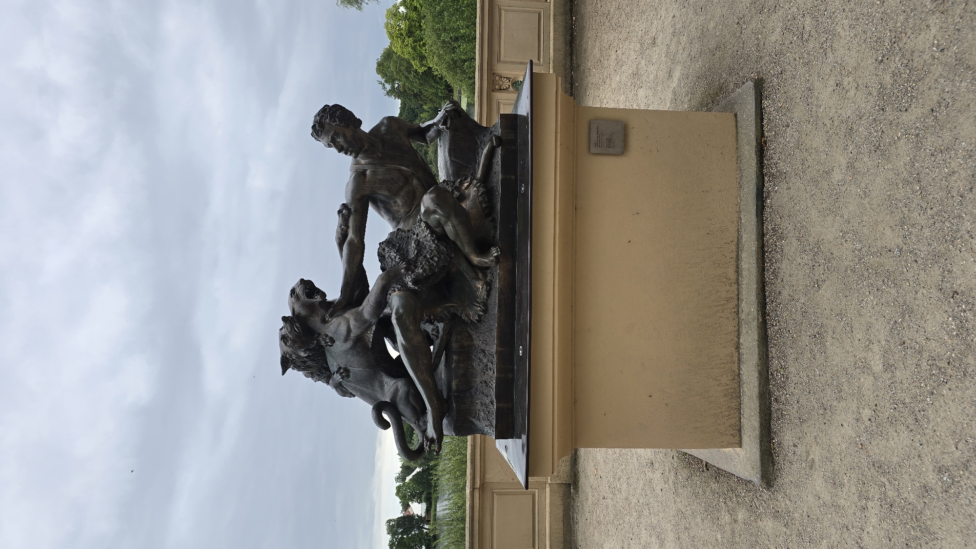 A bronze sculpture depicting a struggle between a man and two lions is displayed outdoors on a pedestal in Schwerin, Germany. The sculpture is positioned on a light brown, rectangular pedestal in what appears to be a park or garden area. The man, with muscular physique, is shown in the center, grappling with one lion that is biting his arm and chest, while another lion is attacking him from behind. Both lions are depicted in dynamic poses, with open jaws and claws extended. The man's facial expression suggests pain and resistance. The sculpture is placed on a dark base, which sits on the light brown pedestal. A small, rectangular plaque is affixed to the front of the pedestal, likely providing information about the artwork. The surrounding environment includes a gravel pathway, a low wall with decorative elements, trees and foliage, and a body of water in the background, possibly a lake. The sky is overcast with gray clouds.