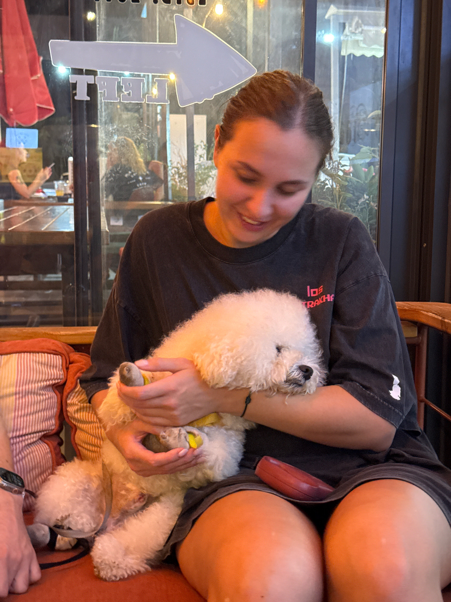 A young woman with brown hair tied back is seated in what appears to be a cafe or restaurant. She is holding a small, fluffy white dog in her arms and smiling. The dog's face is partially obscured by its fur. The woman is wearing a dark gray t-shirt with a pink logo on the left side and a white graphic on the right sleeve. She is sitting on a textured orange and white striped cushion, and her legs are visible. A dark red object, possibly a wallet or phone case, rests on her lap. Behind the woman, a large window reflects light, showing a blurred image of other patrons and possibly outdoor elements. A white arrow graphic is adhered to the glass, pointing to the right, with the word "TELL" visible in white block letters below it. The environment appears to be indoors with lighting that suggests it is evening. The location is identified as Konyaaltı, Turkey.