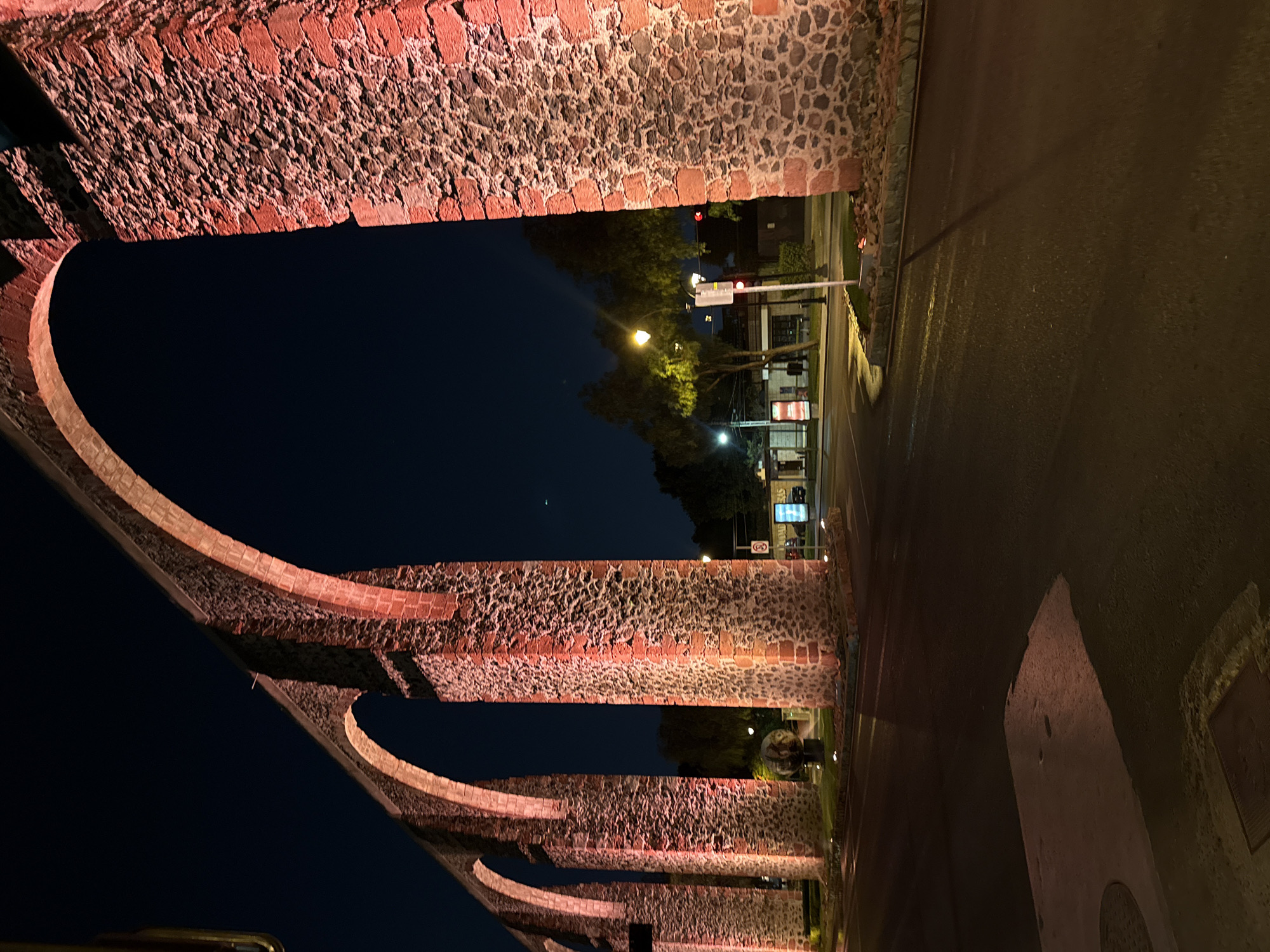 The image shows the aqueduct of Querétaro at night. The aqueduct, constructed of pink stone, spans multiple arches and is illuminated by warm lighting. A street runs alongside the aqueduct, with streetlights, traffic signals, and trees visible. The sky is dark. There are no people visible in the image. The focus is on the architectural structure and the urban landscape at night.