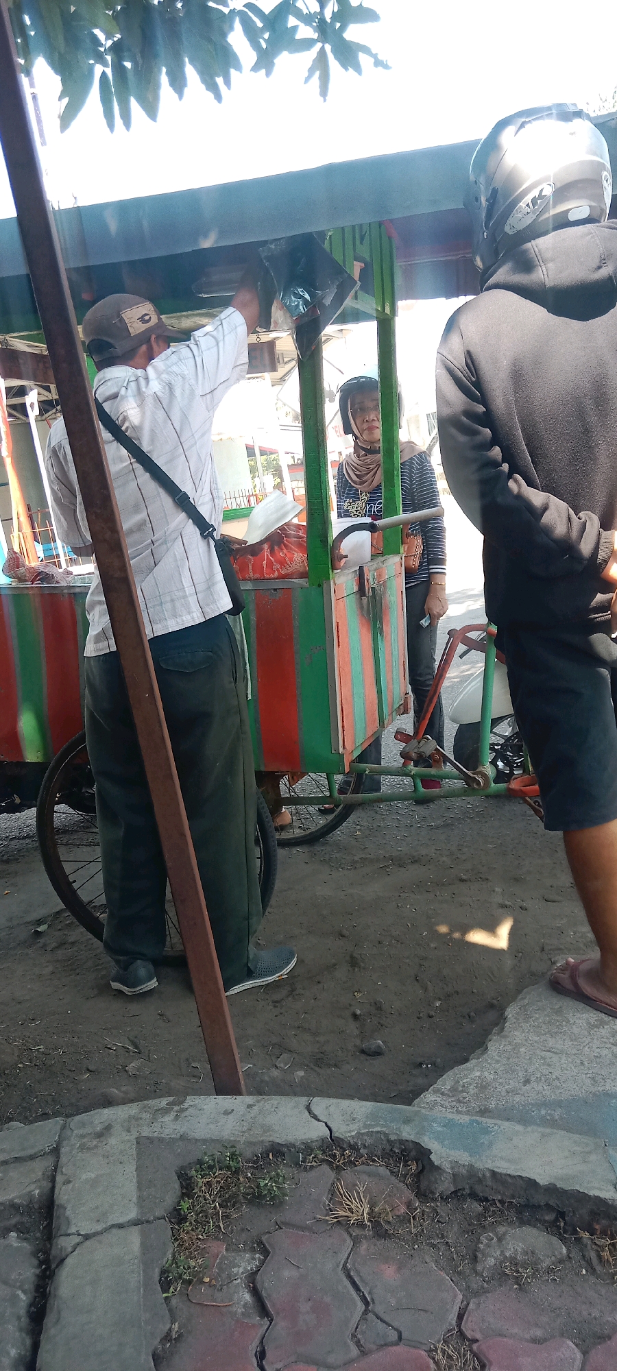 A street vendor is serving a customer from a food cart. The vendor, a man wearing a white plaid shirt, green pants, and a brown cap, is handing a black bag to a customer. The customer, a woman wearing a hijab and a striped shirt, is partially visible behind the cart. A second customer, wearing a black hoodie and a helmet, stands to the right. The food cart is colorful, with red and green stripes. The scene appears to be on a street in Beru, Indonesia, with a tree and sky visible in the background.