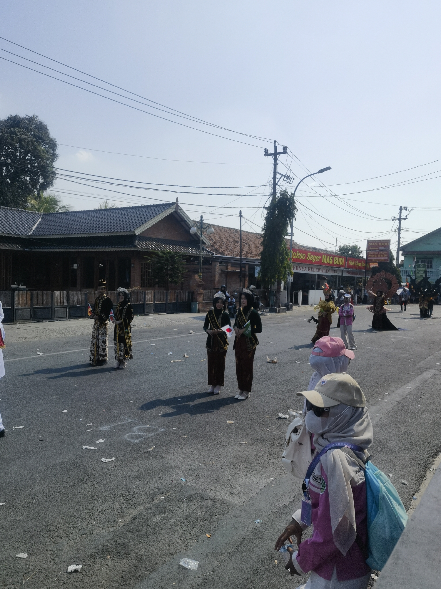 The image captures a street scene in Blora, Indonesia, likely during a parade or cultural event. In the foreground, a person wearing a hijab, a face mask, sunglasses, a pink shirt, and a beige baseball cap is walking, carrying a light-colored bag and a blue backpack. Their left hand is holding a clear plastic bottle. Behind this person, several individuals are participating in what appears to be a procession. Two individuals in traditional attire, a man and a woman, stand side-by-side holding small flags. The woman is wearing a hijab and a crown-like headpiece, and both are dressed in dark patterned clothing with sashes. Further back, other participants are visible, some in elaborate costumes, including one with a large, intricate headdress. Buildings line the street, including one with a red sign that reads "Bakso Seger Mas Budi". Power lines crisscross the sky, and streetlights are visible. The road surface is asphalt, with some debris and white markings visible. The overall atmosphere suggests a public event under a clear, sunny sky.
