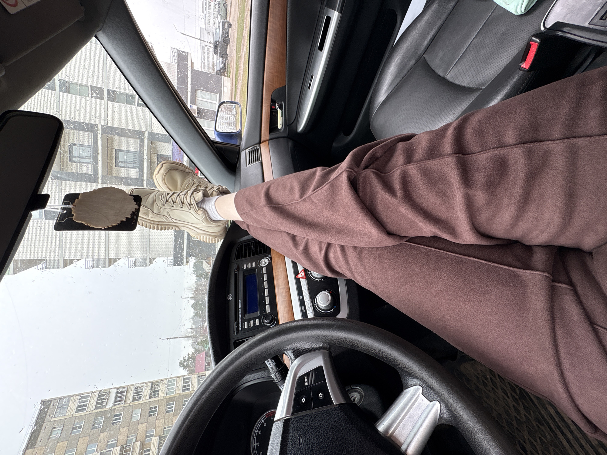 A view from inside a car reveals a person with their feet propped on the dashboard, obscuring the view of the exterior. The individual wears brown sweatpants and light-colored sneakers with white socks. Outside the windshield, buildings suggest an urban environment, and rain streaks the glass. The car's interior features a black dashboard with woodgrain trim. The steering wheel is in the foreground. An air freshener hangs from the rearview mirror. The city is Chaikovsky, Russia.