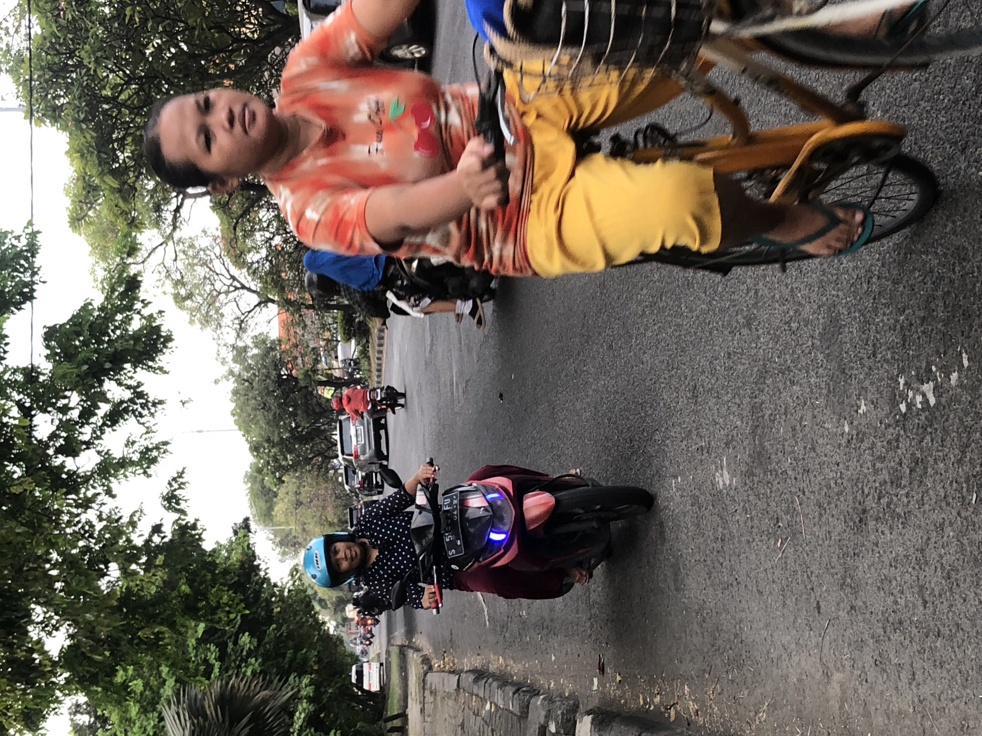 The image captures a street scene in Sumorame, Indonesia, featuring two women navigating the asphalt road. In the foreground, a woman on a bicycle, wearing bright yellow pants and an orange tie-dye shirt with cherry graphics, is seen from the chest down. Her right foot, clad in a teal flip-flop, is pushing the pedal. The bicycle has a black basket in front, partially obscuring the lower half of her body. Behind her, to the left, another woman rides a pink and black scooter. She is wearing a dark polka dot shirt, maroon pants, and a light blue helmet. The scooter has illuminated blue lights at the front. Both women appear to be moving forward, engaged in their respective modes of transport. The environment is urban, with a tree-lined street and other vehicles, including cars and motorbikes, visible in the background, suggesting moderate traffic. The road surface is asphalt, with some scattered debris.