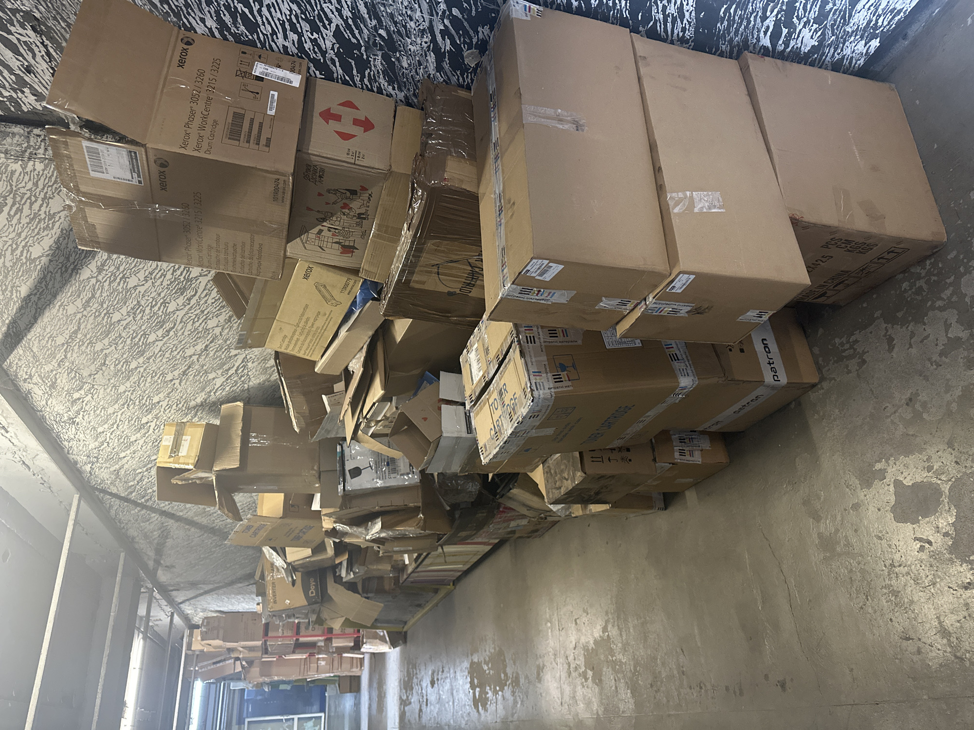 The image shows a chaotic pile of cardboard boxes stacked against a wall in what appears to be an indoor storage area or warehouse. There is a mix of boxes of various sizes and types, with some showing the brand "Xerox" along with product names like "Phaser 3052/3260" and "WorkCentre 3215/3225", indicating they contain printer-related components such as drum cartridges. Other boxes display terms like "TONER CARTRIDGE" and "INVERTER SOLAR". Some boxes appear to be slightly crushed or damaged. The floor is concrete and shows signs of wear, with an uneven surface. The location seems to be a storage or transitional area for discarded or stored items, possibly within an industrial or commercial setting in the city of Unknown, Unknown. No people are visible in the image.