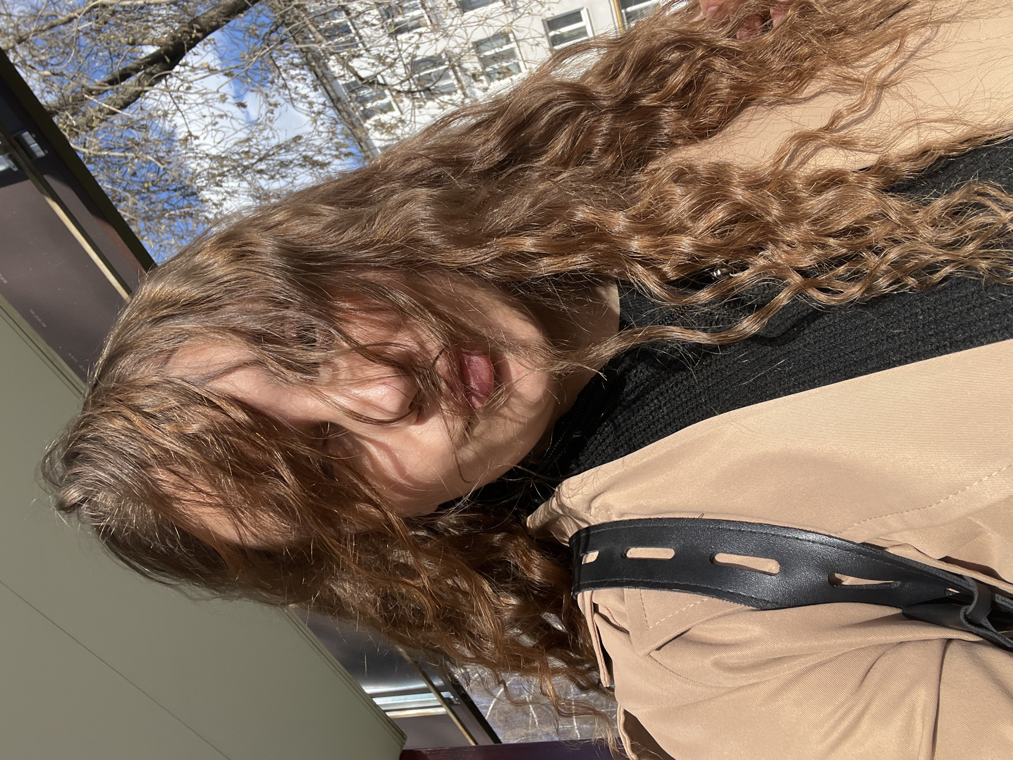 A woman with long, curly brown hair is captured in a close-up outdoor shot. She is wearing a black sweater and a beige jacket with a black strap detail. The background reveals a city environment, with a building with windows visible, along with trees and a glimpse of a partly cloudy sky. The specific location is Chaikovsky, Russia, as requested. The image is taken at a slightly tilted angle, obscuring the woman's face with her hair.