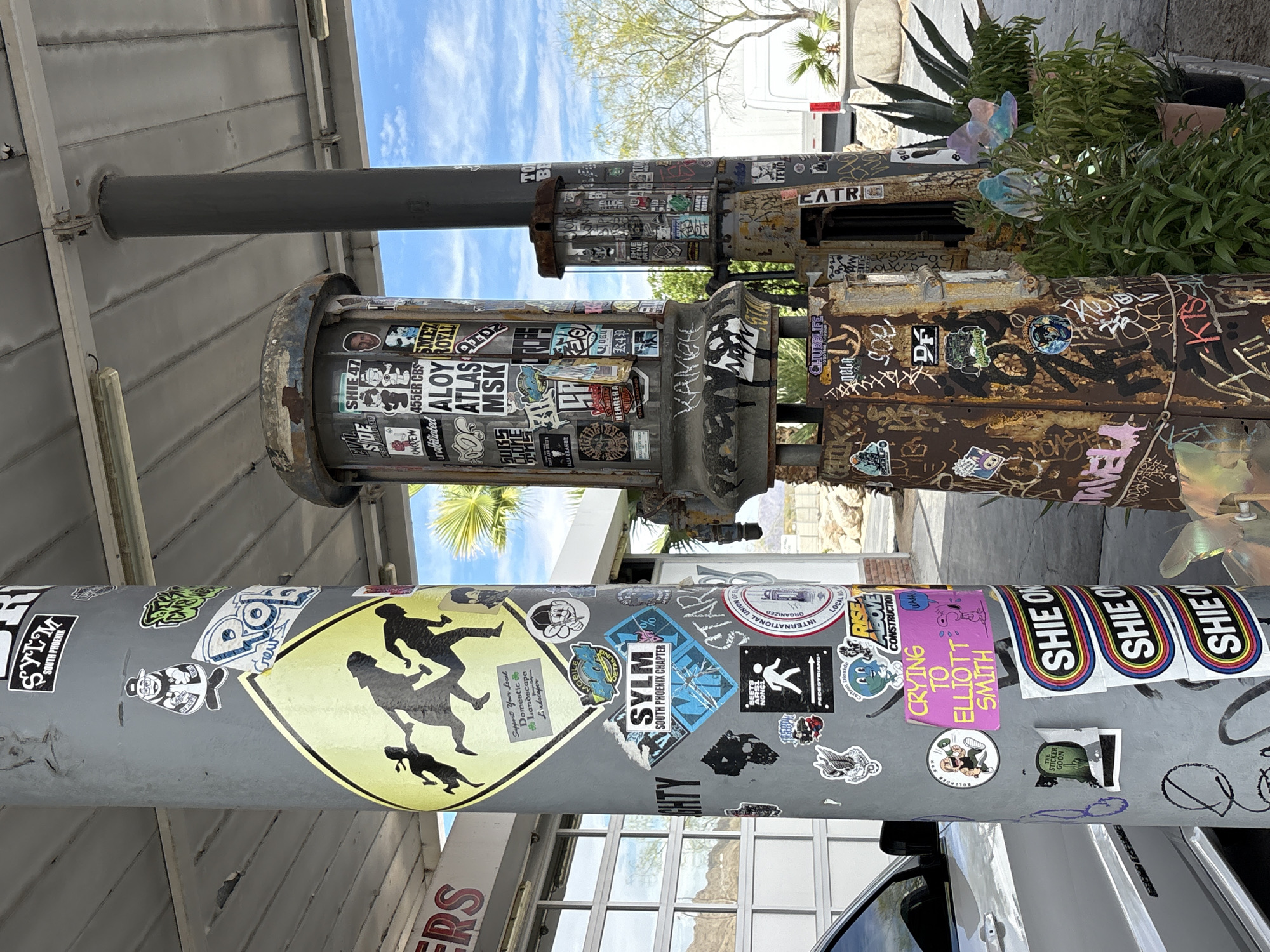 The image shows a close-up view of a utility pole covered in stickers, with two old gas pumps behind it. The pole on the left is grey and adorned with numerous stickers of various sizes and designs, including a large yellow diamond-shaped sticker with silhouettes of people running. The gas pumps on the right are rusted and also covered in graffiti and stickers. Behind the pole and pumps, a white building with large windows and a covered walkway is visible. A silver car is parked to the left of the pole. In the background, there are some green plants, including an agave plant, and a blue sky with some clouds. The scene appears to be outdoors, possibly at a gas station or a similar public area in Chiriaco Summit, United States, given the desert-like vegetation and dry climate suggested by the surroundings.