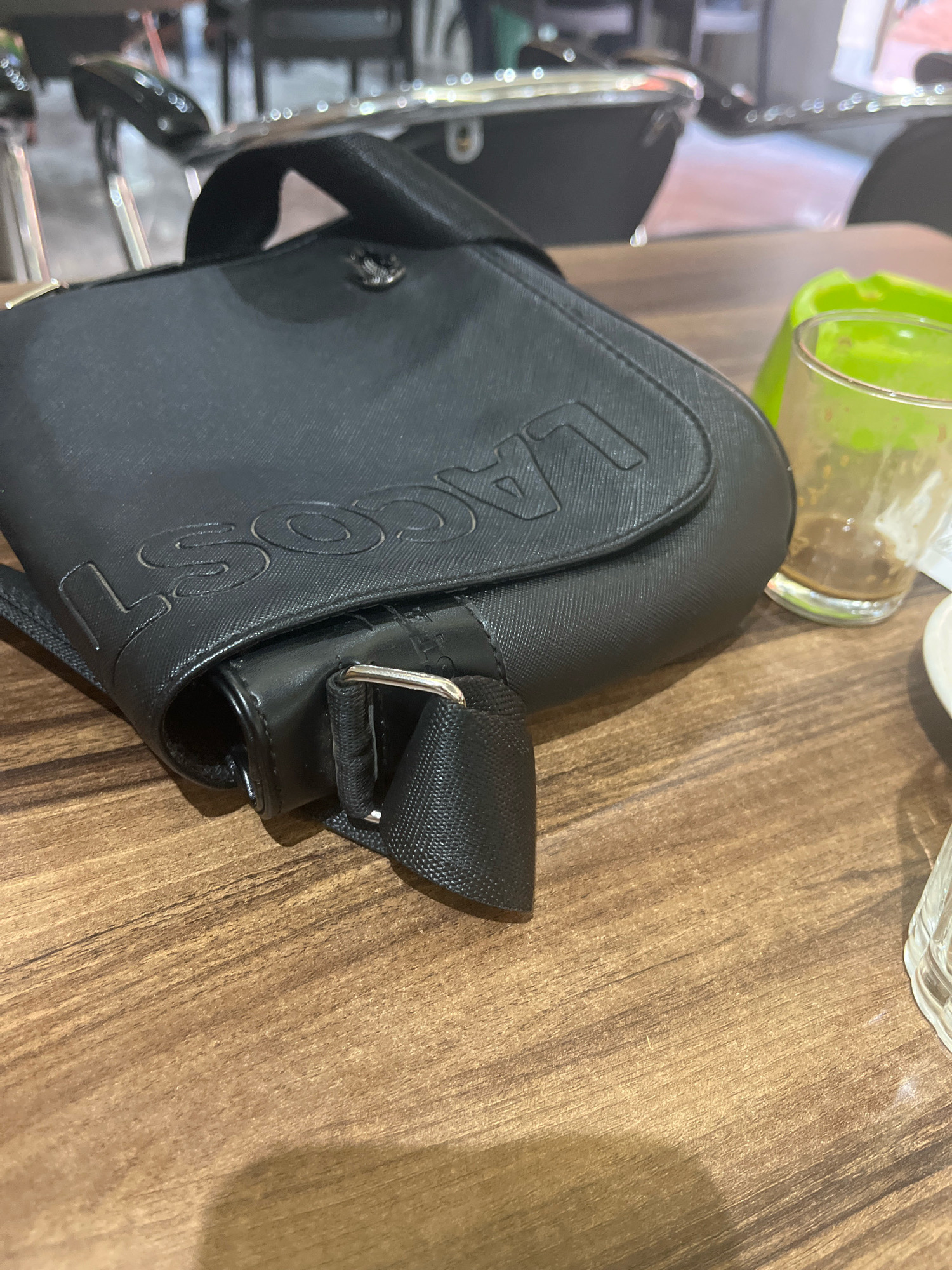 The image depicts a close-up view of a black, textured messenger bag with "LACOSTE" embossed on its flap, resting on a wooden surface. A clear glass with a yellowish residue and a green object, possibly a lime wedge or plastic, are visible to the right of the bag. The background is blurred, showing hints of furniture, likely chairs, and a metallic structure. The scene appears to be indoors, possibly in a café or restaurant. There are no people visible in the image, and no specific details suggest a location in Azilal, Morocco.