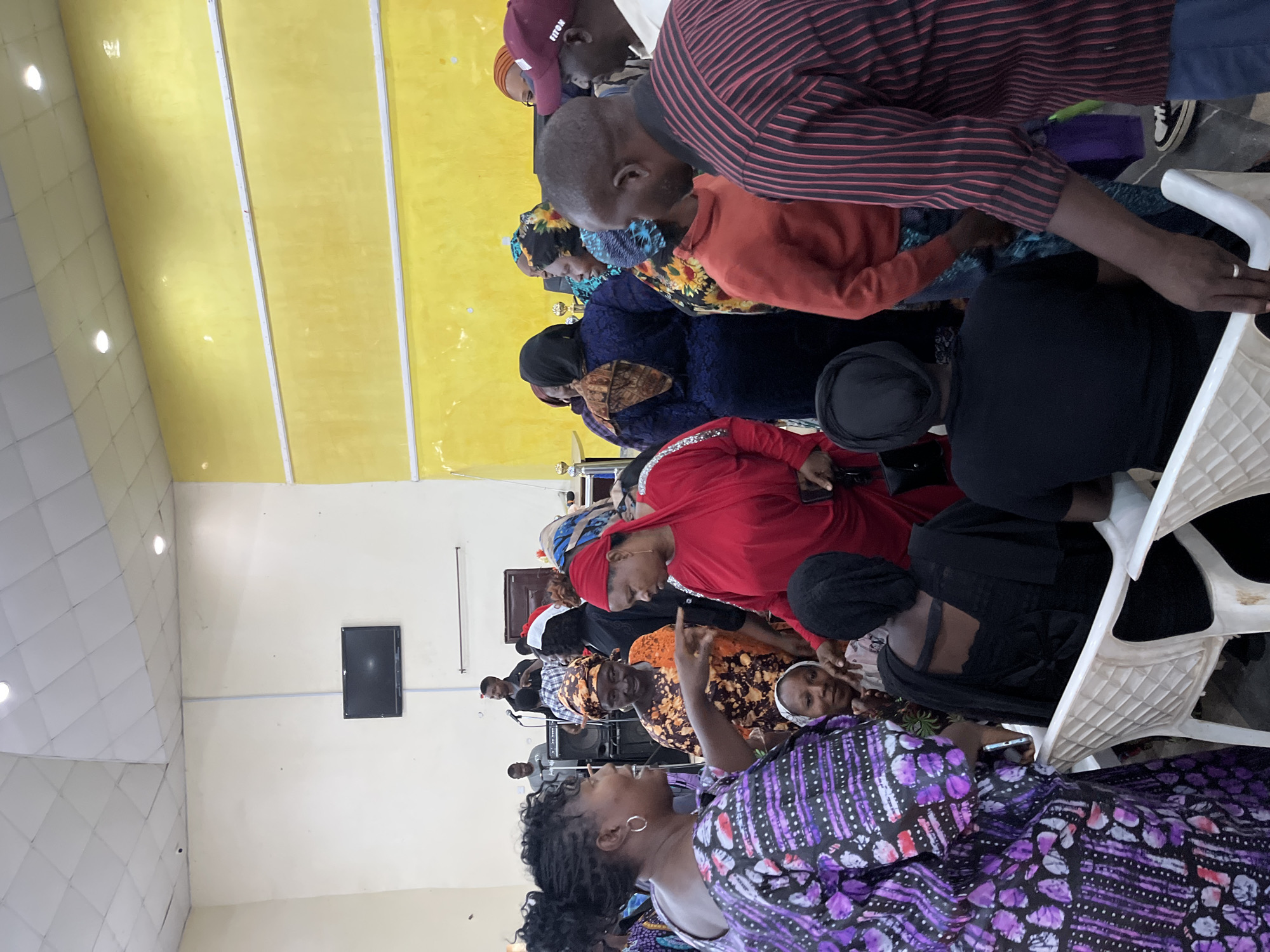 A photograph shows a group of people inside a room. The room has yellow walls. The people are seated in chairs and wearing a variety of clothing. A woman in a purple patterned dress is standing and talking to the group. A television is on the wall. The location is likely Jos, Nigeria, due to the clothing and the people pictured.