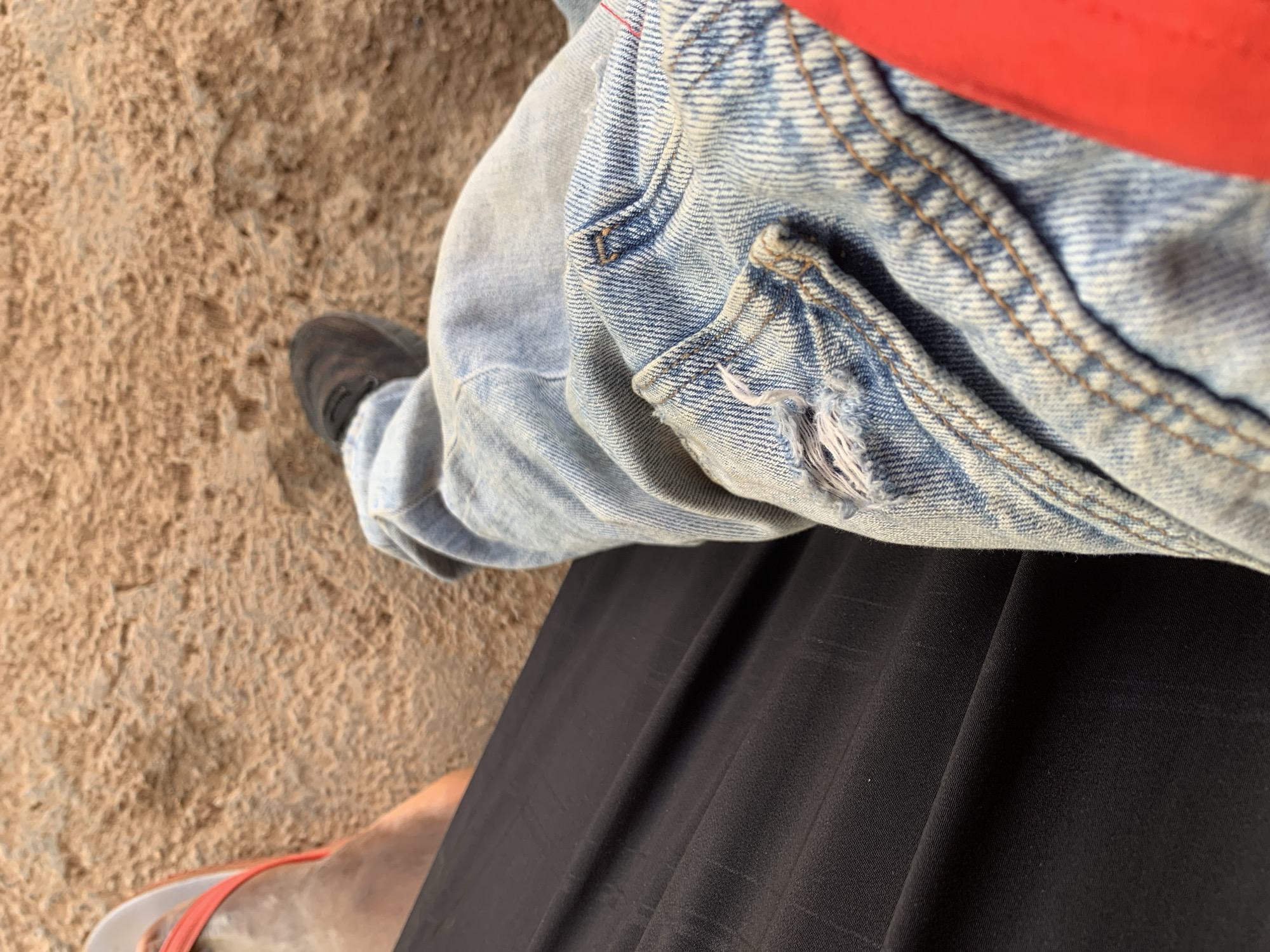 The image is a close-up shot of a person's legs, seen from a first-person perspective. The person is wearing light blue denim jeans with visible distressing and a black fabric item, possibly a skirt or pants. On the left side of the frame, a bare foot with a bright orange flip-flop is visible, resting on a sandy surface. In the background, also sandy, a dark-colored shoe is visible. The overall scene suggests a casual outdoor setting. The sandy environment could indicate a location like the Rwang Pam Township Stadium area in Jos, Nigeria, known for its sandy pitches and surrounding dry terrain.