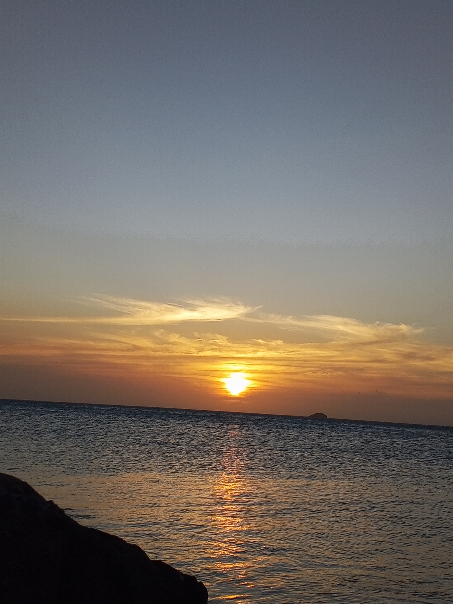 The image depicts a sunset over the ocean in Pedro González, Venezuela. The sky transitions from a light blue at the top to shades of orange and yellow near the horizon. There are thin, wispy clouds near the sun. The sun is setting directly over the water, with a reflection stretching towards the foreground. The ocean is relatively calm, with small waves visible. A dark, rocky outcropping is visible in the bottom left corner. A small island is visible on the horizon to the right of the sun.
