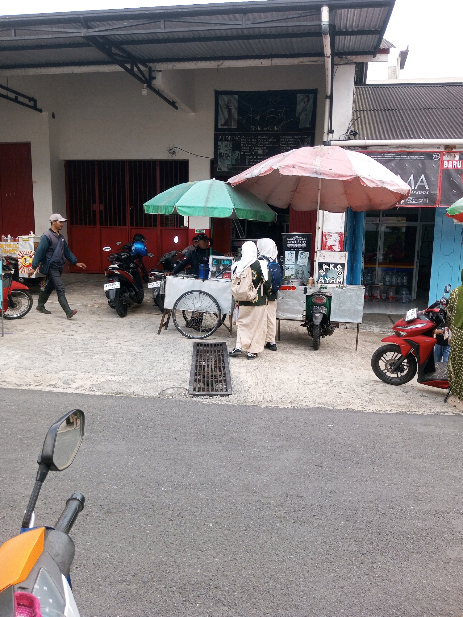 The image depicts a street scene in Krasak, Indonesia, near a building with a corrugated metal roof and a white facade. In front of the building, there is a food cart with a large, open pink umbrella. Two young women in light-colored uniforms and headscarves are standing at the food cart, looking at the items on display. A man wearing a light-colored cap and dark clothing walks by the cart. Several motorcycles are parked along the street. One red motorcycle is parked in the foreground on the right side of the image, and another red motorcycle is parked on the left. Two black motorcycles are parked behind the food cart. A drainage grate is visible on the ground in front of the food cart. The area appears to be a commercial or street-side vendor location.
