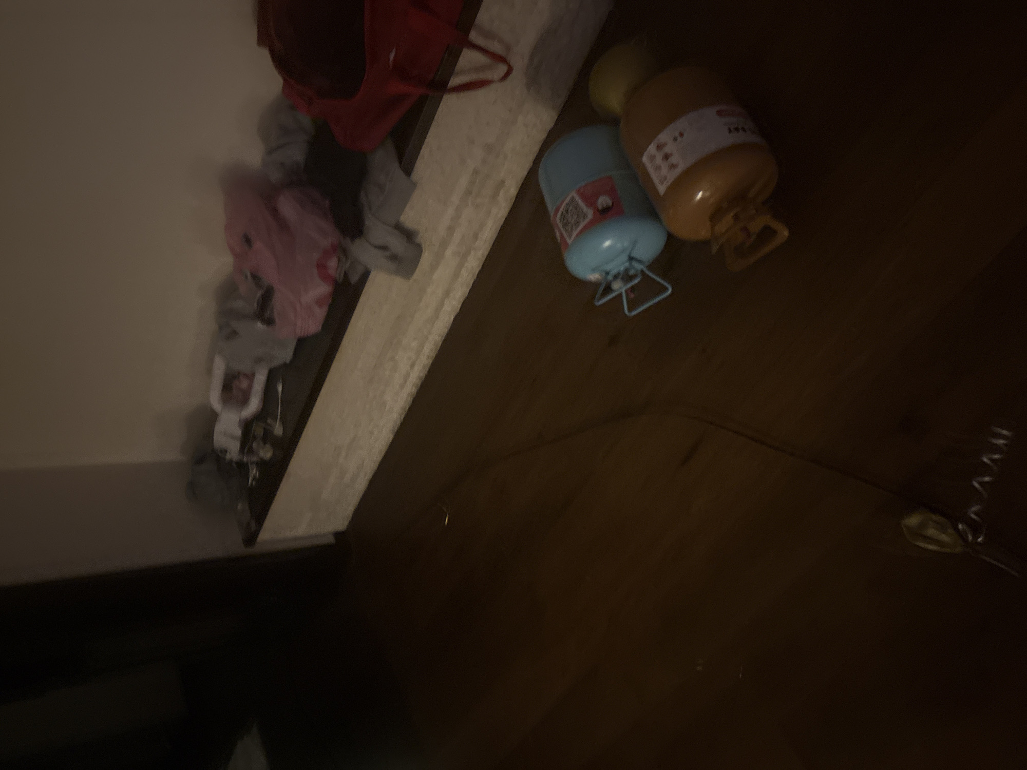 The image depicts an interior scene, likely a room, with a wooden floor and a white, built-in shelf or counter along the wall. On the shelf, several items of clothing are piled, including a pink garment that appears to be a stuffed toy or bag, and some gray clothing. To the right of the clothing, a red bag is visible. On the floor in the foreground, there are two cylindrical objects, possibly canisters or tanks, one blue and one orange. The blue object has a handle and labels with text and a QR code. The orange object also has labels. The room is dimly lit, creating shadows and a somewhat cluttered appearance. The location is stated to be in the city of Orekhovka, Russia, but there are no specific visual cues in the image to confirm this geographical location. There are no people visible in the image, nor any actions or interactions taking place.