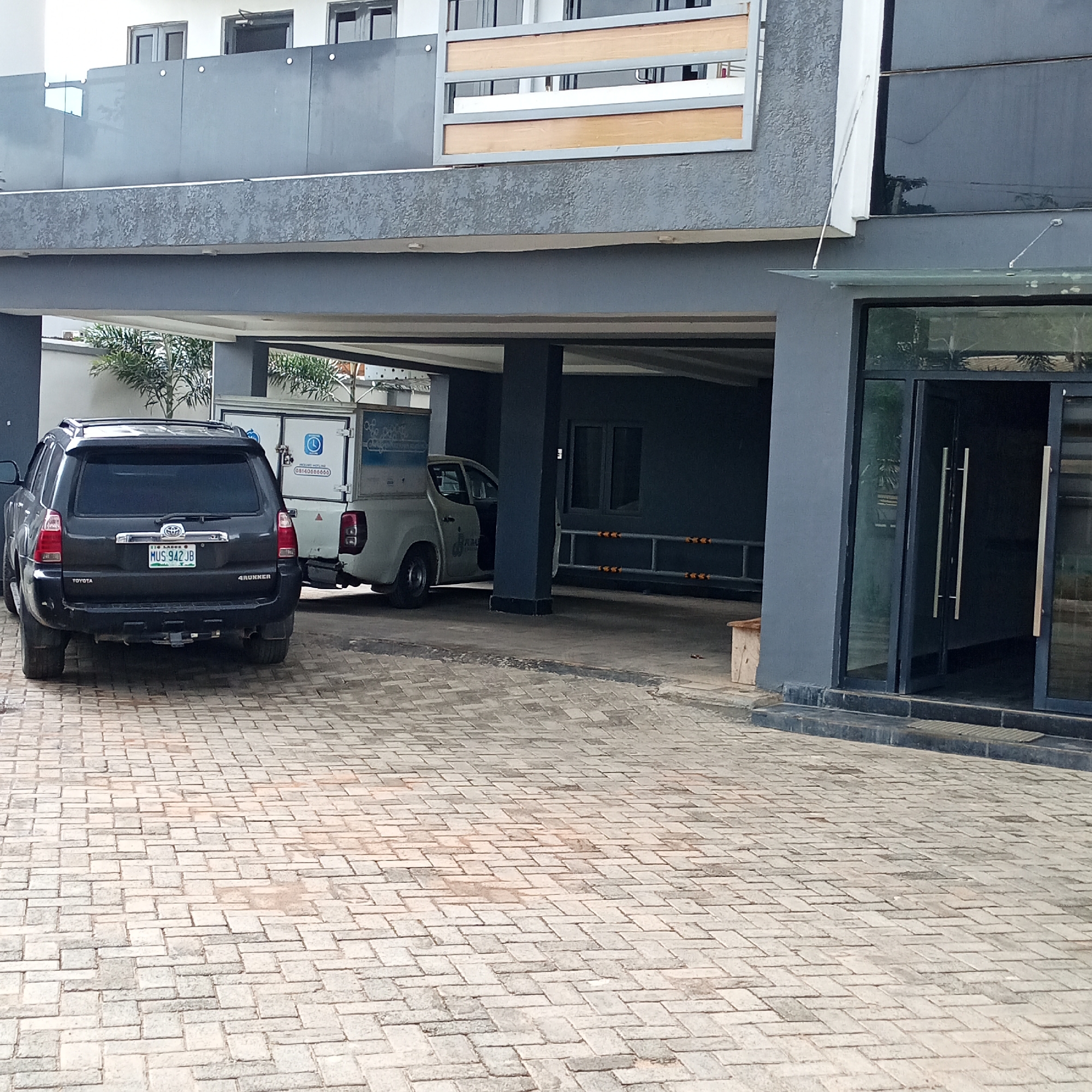 The image depicts a paved driveway in front of a modern building with a dark gray facade and a glass entrance. Two vehicles are parked on the driveway: a dark-colored Toyota 4Runner SUV in the foreground and a white pickup truck with a utility box in the background. The SUV has the license plate "MUS 942 JB". Palm trees and other greenery are visible behind the vehicles, suggesting a tropical or subtropical climate. The building features large windows and multiple levels, with a balcony or overhang on the second floor. The sky is not visible, but the lighting suggests it is daytime. The scene appears to be a private or commercial property in Ibadan, Nigeria. No people are visible, and the overall atmosphere is still and quiet. The paving stones are laid in a herringbone pattern. Visible text includes "TOYOTA" and "4RUNNER" on the SUV, and lettering on the utility box of the white truck which includes a phone number "08140666666".