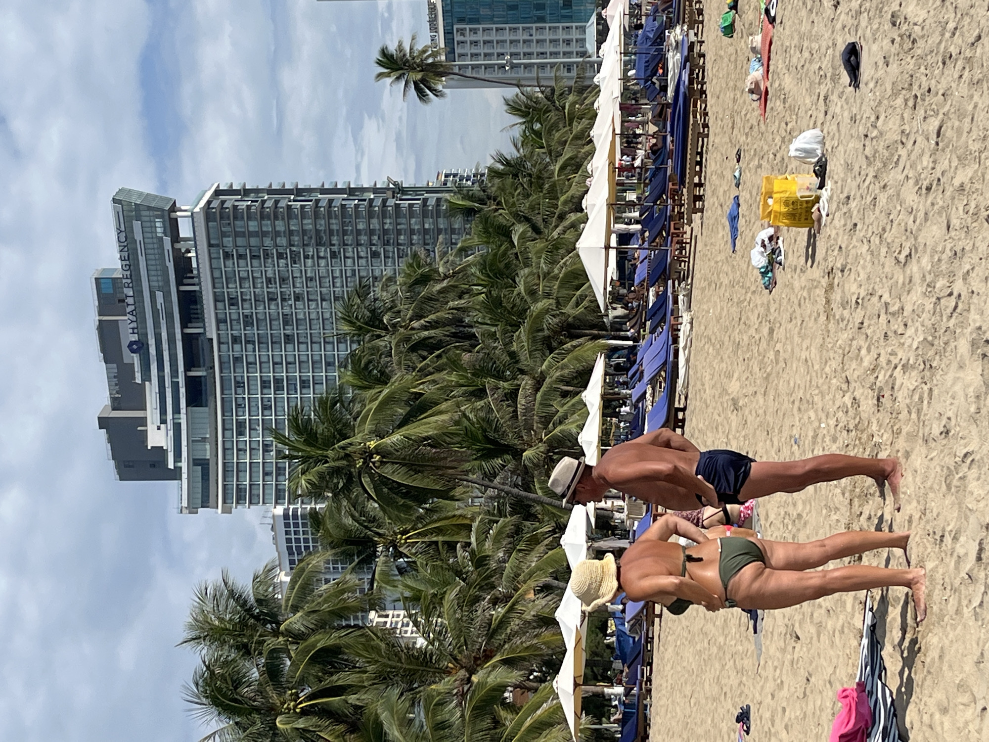 The image shows a beach scene in Nha Trang, Vietnam, with a prominent Hyatt Regency hotel in the background. In the foreground, a man wearing a straw hat and dark swim trunks stands on the sand, facing away from the camera. Next to him, a woman in a straw hat and a green bikini is also facing away, with her left hand on her hip. The beach is lined with rows of blue lounge chairs and white umbrellas. Scattered on the sand are various personal items, including towels, bags, and flip-flops. Palm trees are visible throughout the scene, adding to the tropical atmosphere. The sky is partly cloudy.