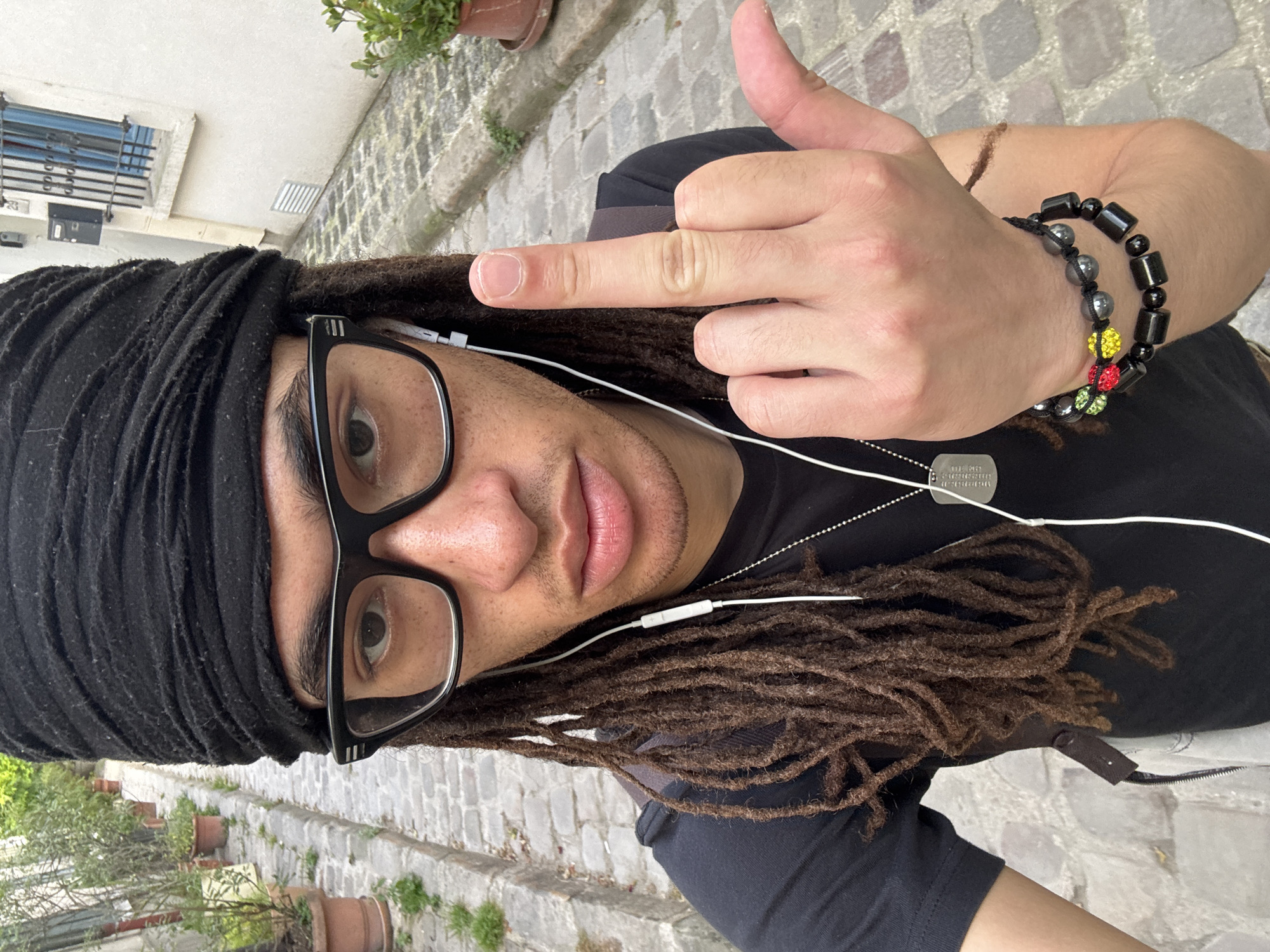 A medium, close-up eye-level shot depicts a Black man wearing a black head wrap, black glasses, and a black shirt, with light brown dreadlocks, facing the camera while giving the middle finger. He wears white earphones and a silver dog tag necklace. He has a beaded bracelet with black, silver, red, yellow and green beads. The background shows a cobblestone street with terracotta pots holding plants on either side. Buildings with white walls are partially visible, and the scene appears to be in an urban environment, possibly in the city of Paris, France.