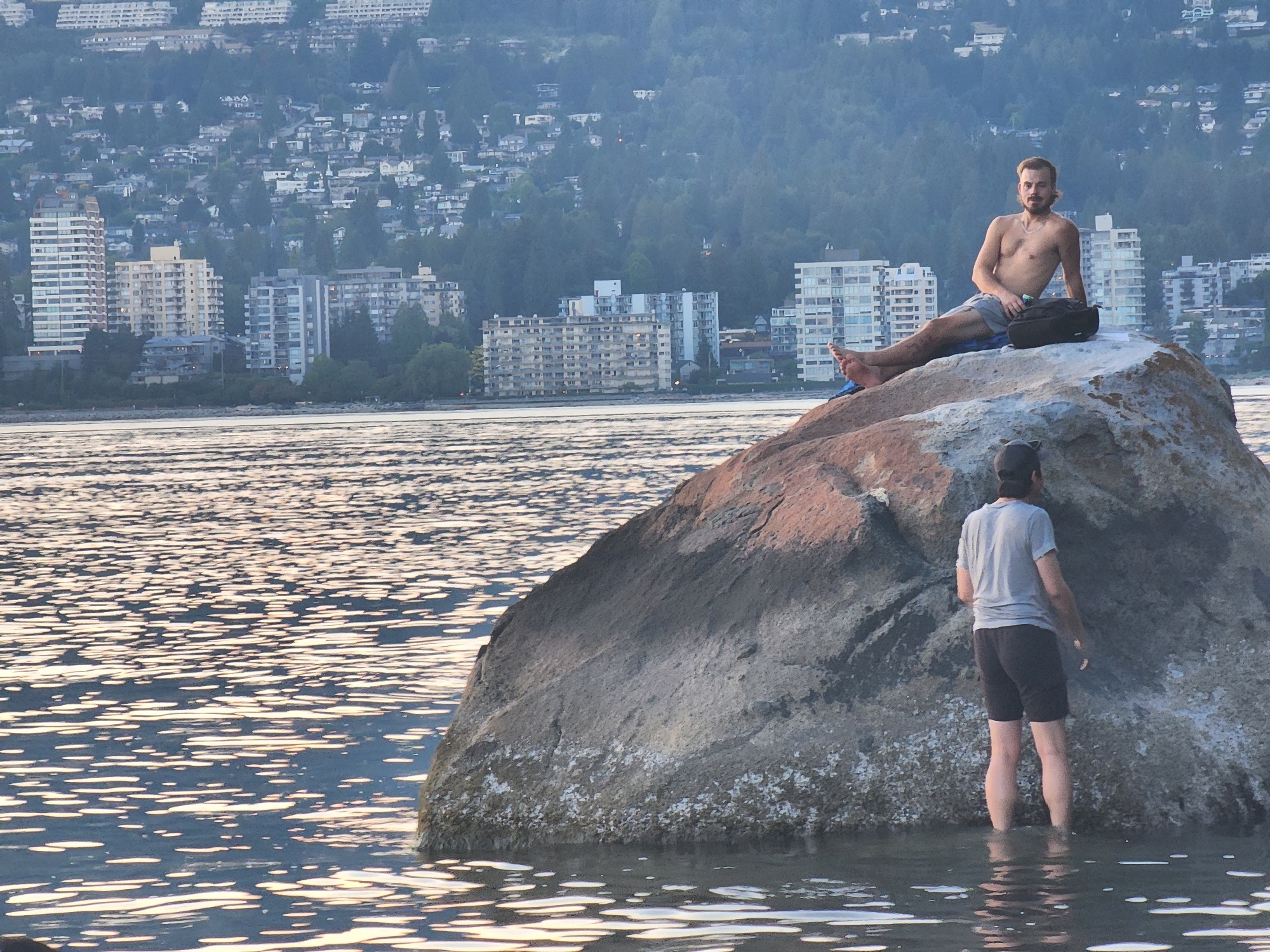 In this media file, two individuals are present on a large rock in the water, with a city skyline in the background. The scene appears to be during the late afternoon or early evening, as evidenced by the soft, warm lighting reflecting on the water's surface.
The first subject, a man, is sitting on the rock, shirtless and wearing grey shorts. He is looking towards the camera with a relaxed expression. Next to him is a black bag. The second subject, also a man, is standing in the water near the rock, facing away from the camera. He is wearing a grey t-shirt and dark shorts, with a baseball cap on. He appears to be wading in the water.
The background features a densely populated city built on a hillside, with numerous buildings ranging from apartment complexes to smaller houses. Lush green trees cover the hills, indicating a natural and developed environment. The water is calm, with ripples creating patterns of light and shadow. The overall atmosphere is peaceful and serene. There is no visible text in the image.